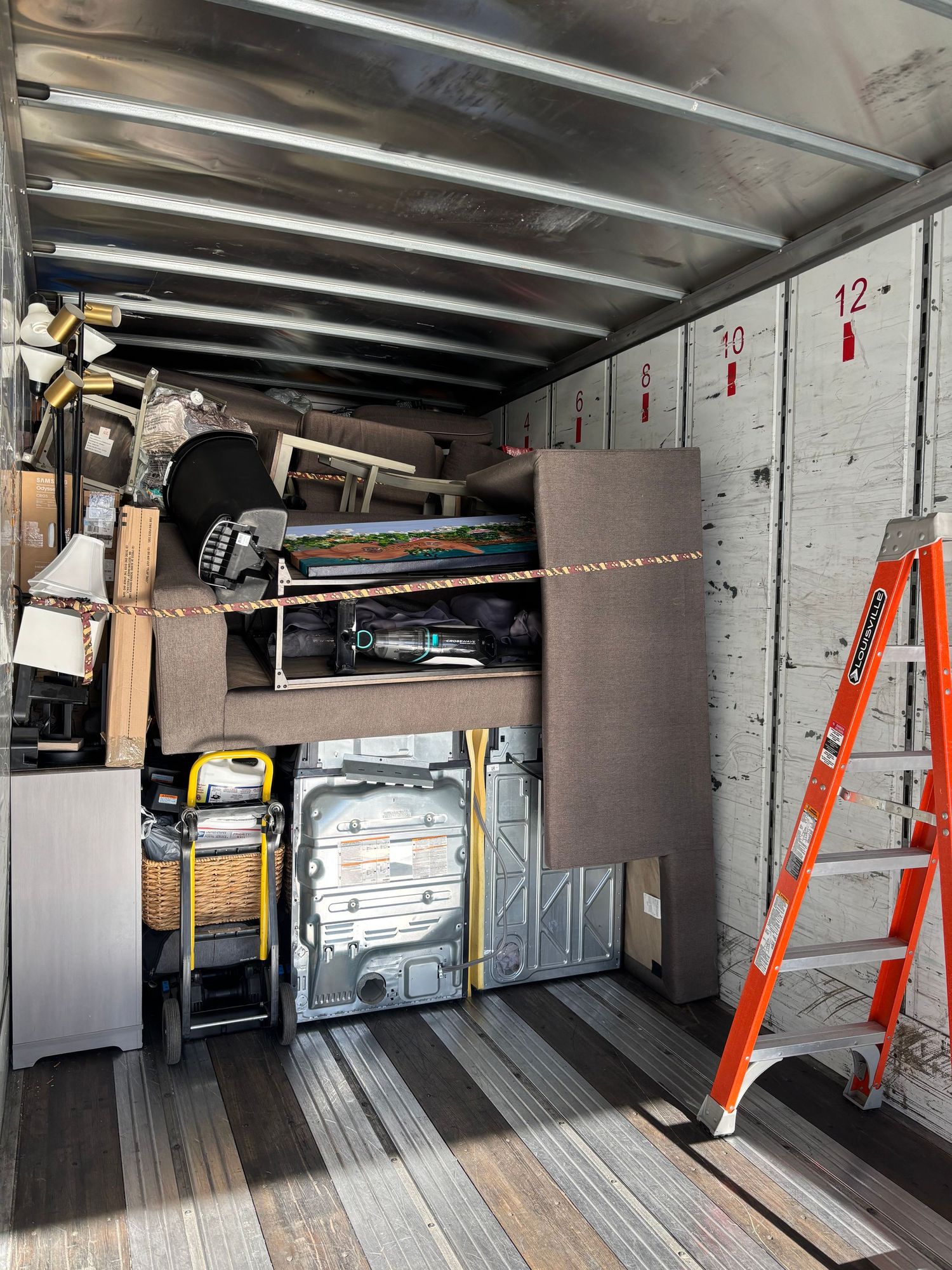 Interior of a loaded moving truck with furniture, appliances, lamps, and an orange ladder secured inside.