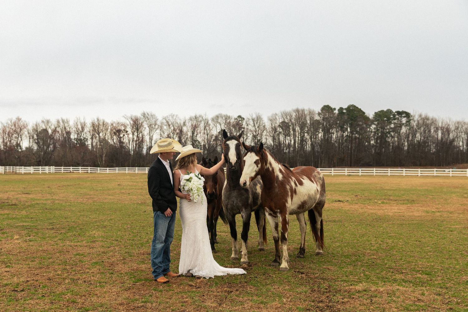 Bride and groom in cowboy hat pose horses in a green pasture on their rustic country wedding day in North Carolina