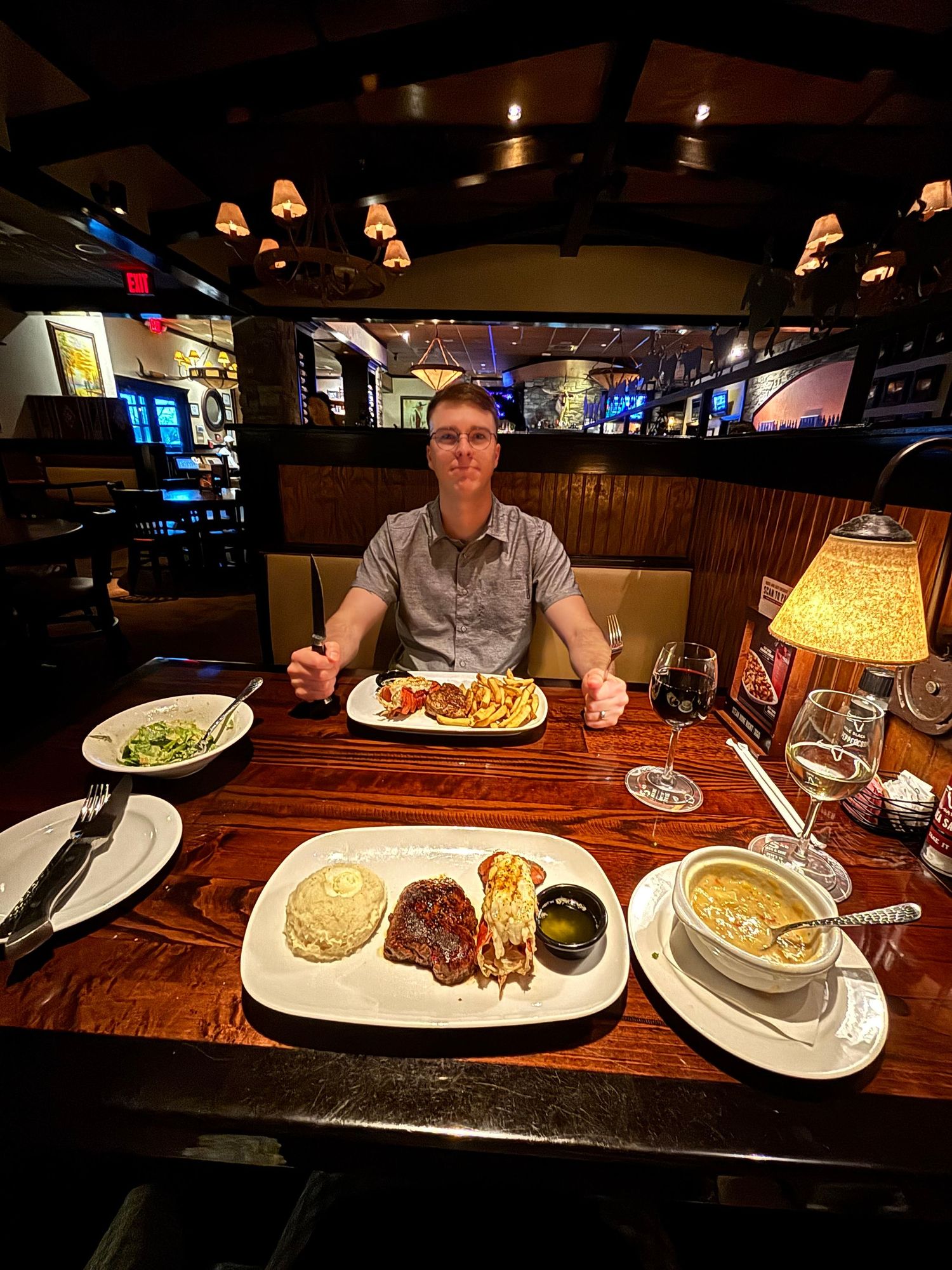 Man dining at upscale steakhouse with surf and turf meal, lobster, steak, soup, and sides on wooden table.