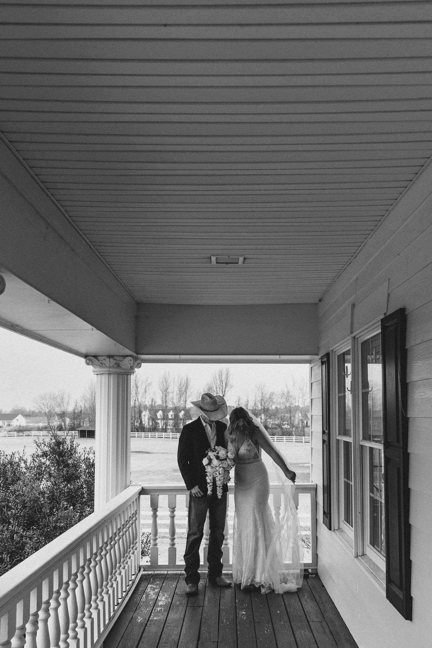 Black and white photo of a couple in wedding attire kissing on a rustic porch, groom in cowboy hat holding bride.