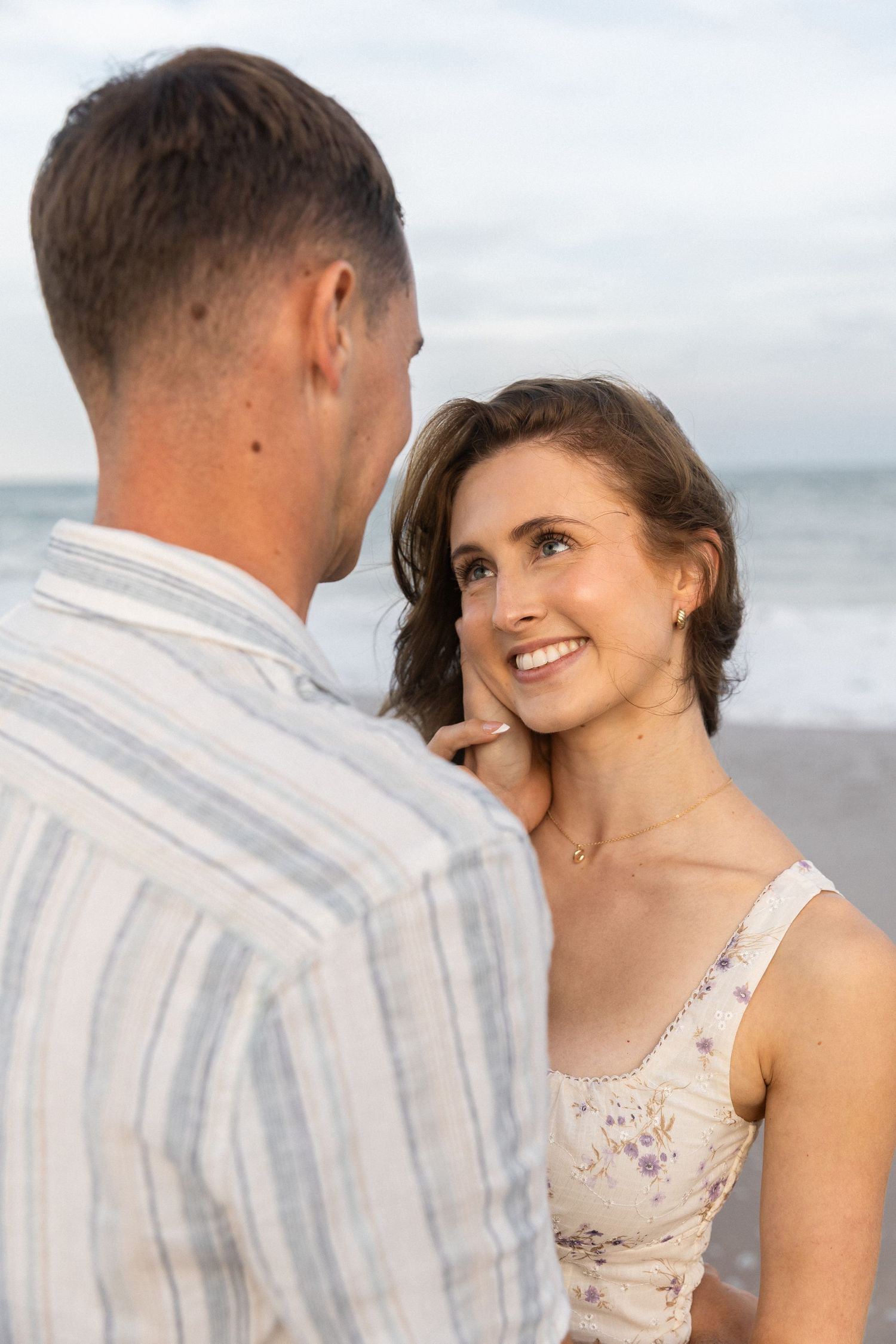 A smiling woman in a floral dress gazes at a man on a beach, with ocean waves in the background.