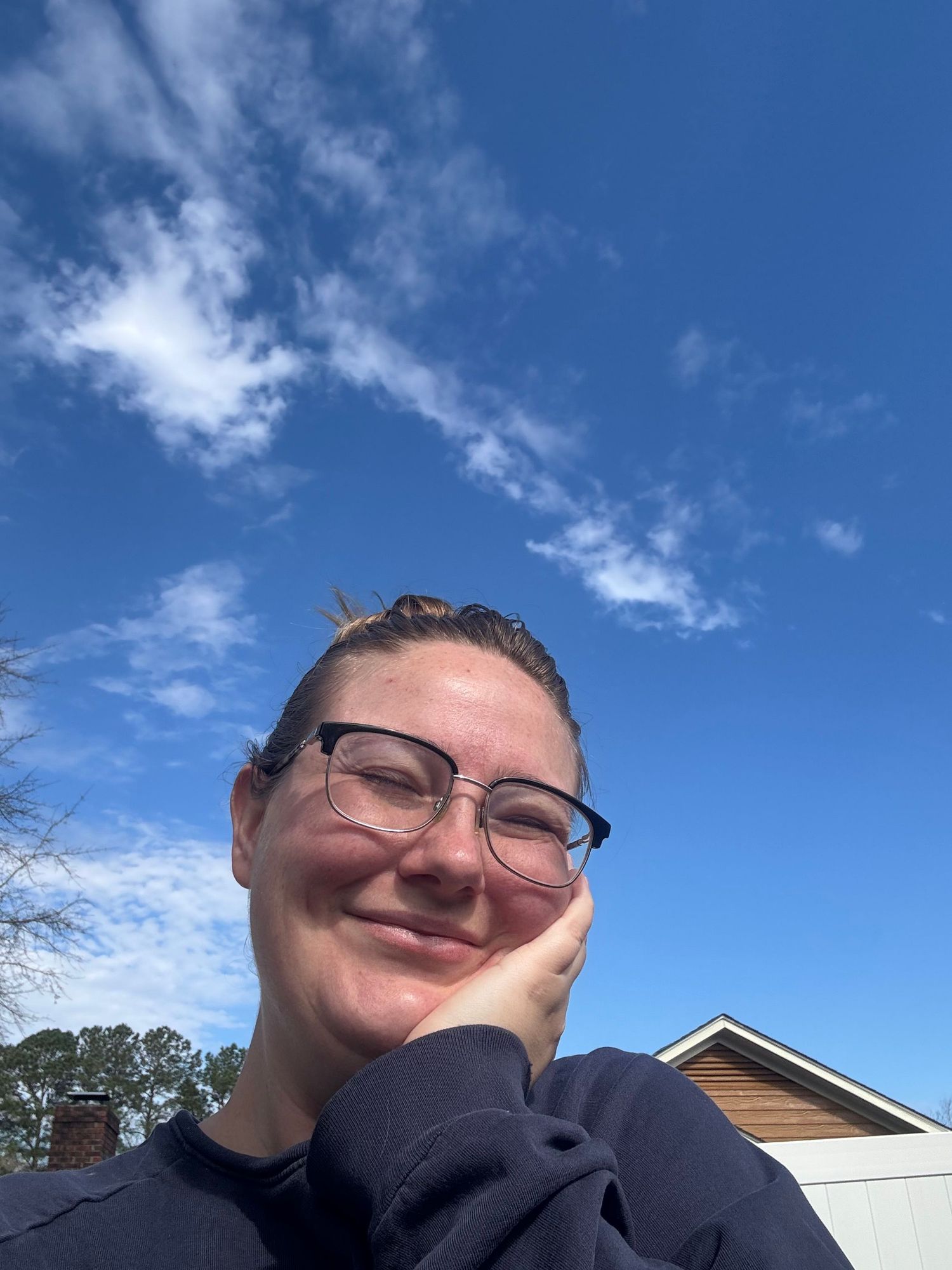 Person with glasses smiling outdoors under a bright blue sky with wispy clouds near a residential building.