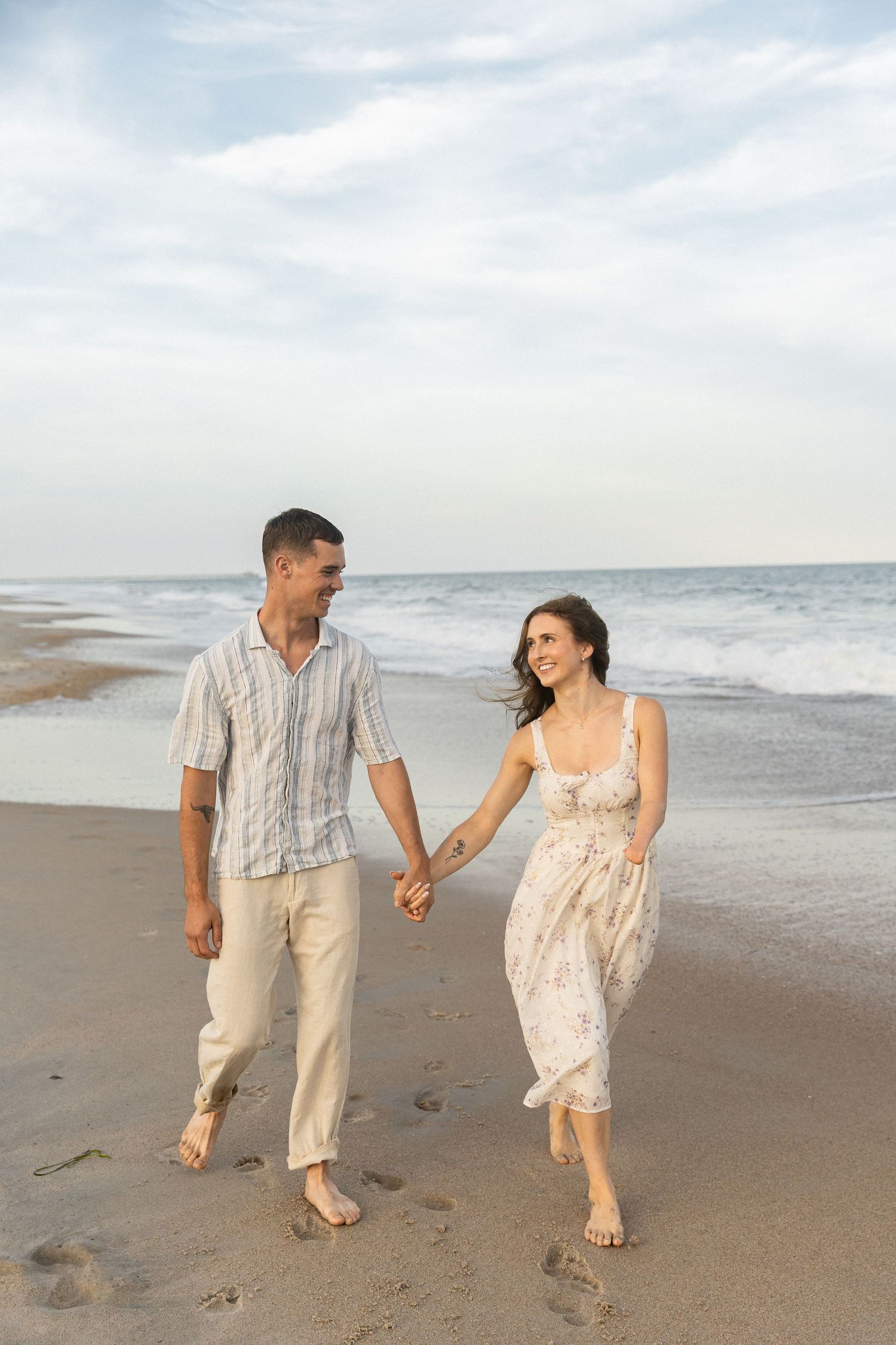 A happy couple holds hands while walking barefoot on a sandy beach with ocean waves in the background.