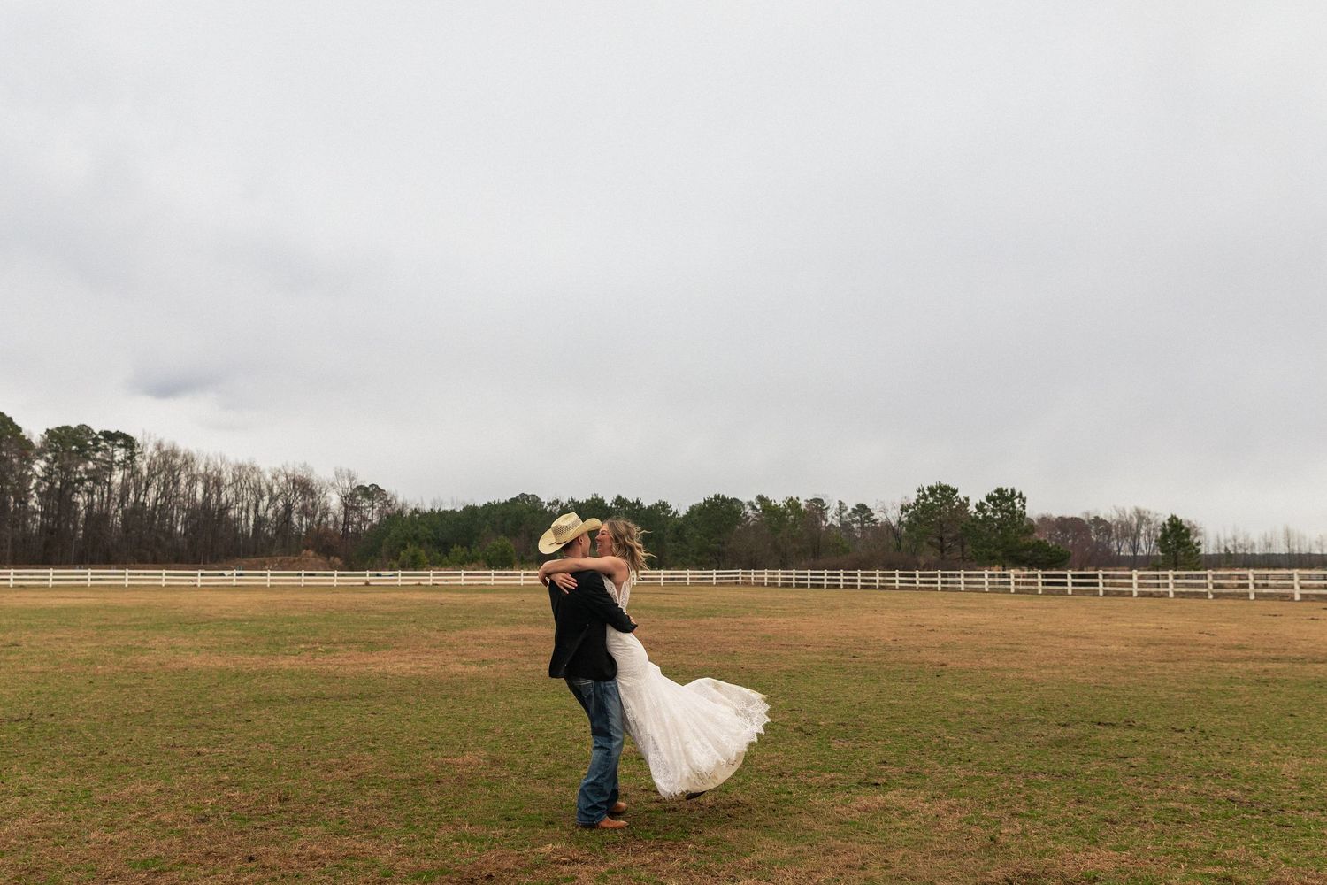Groom lifting bride in wedding dress on a green farm field with white fence and trees in background.
