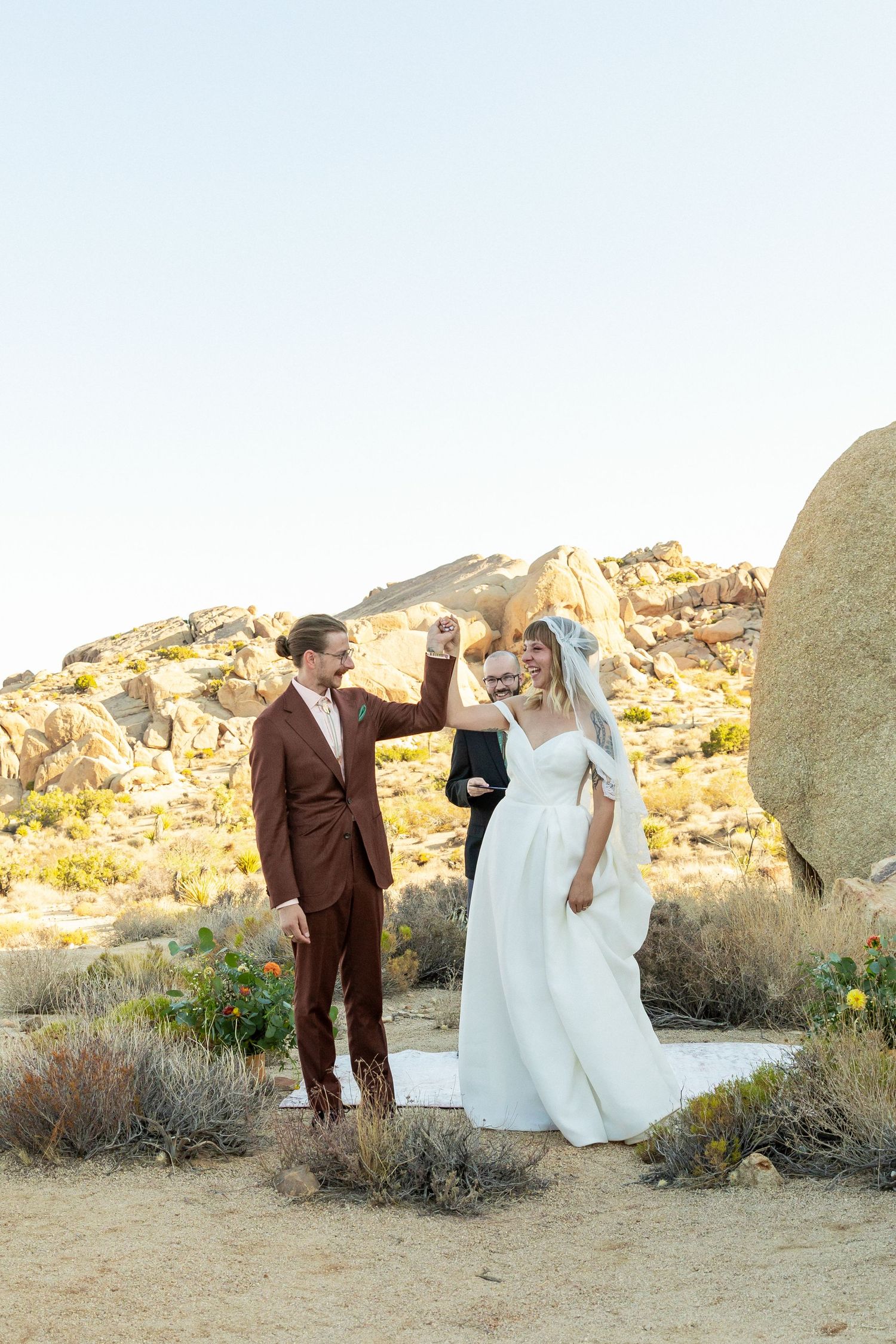 Bride and groom dancing outdoors in desert landscape with rocky boulders, officiant behind them, sunny sky.