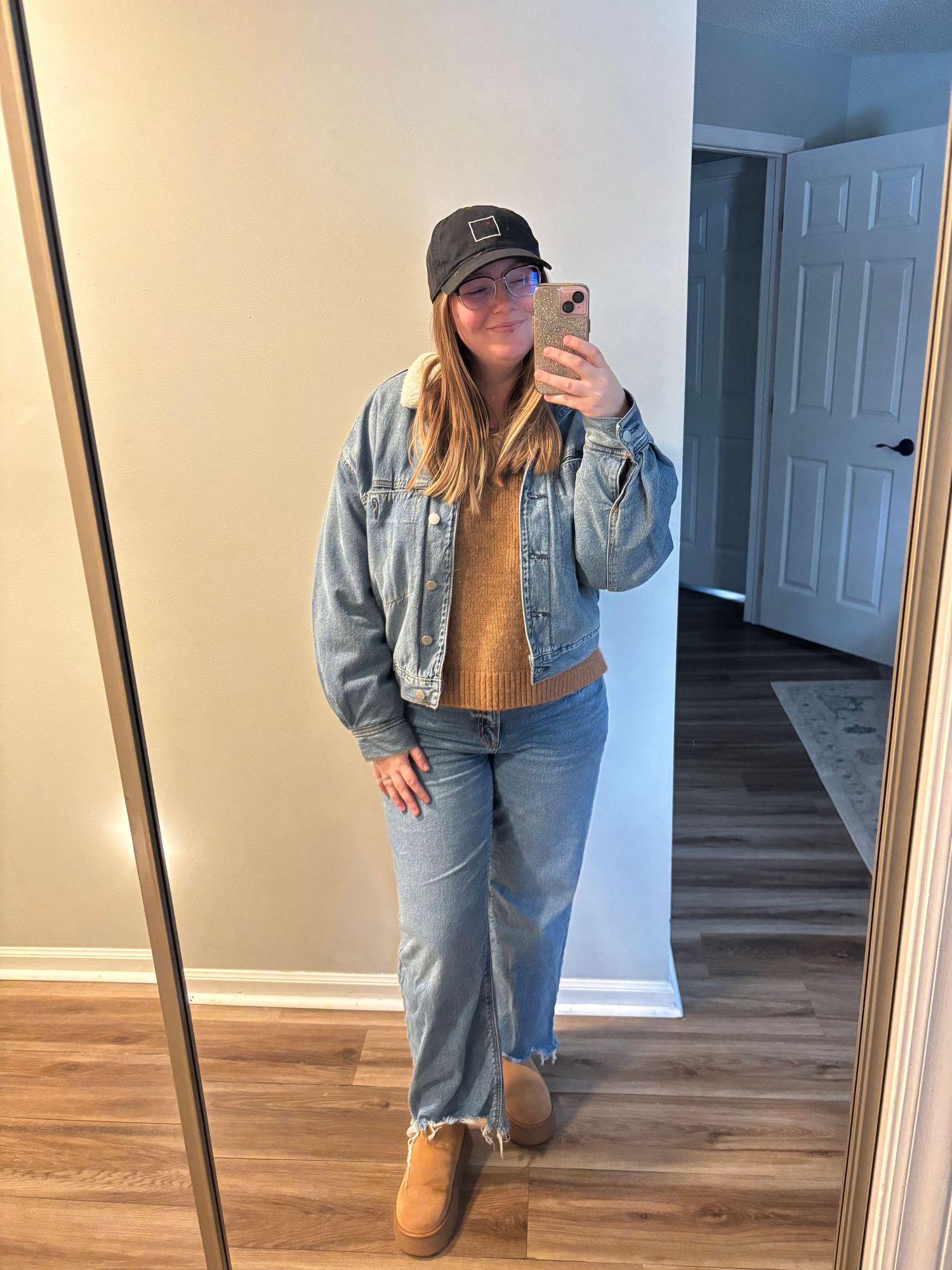 Woman in denim jacket, wide-leg jeans, tan sweater, UGG boots, and black cap takes mirror selfie.