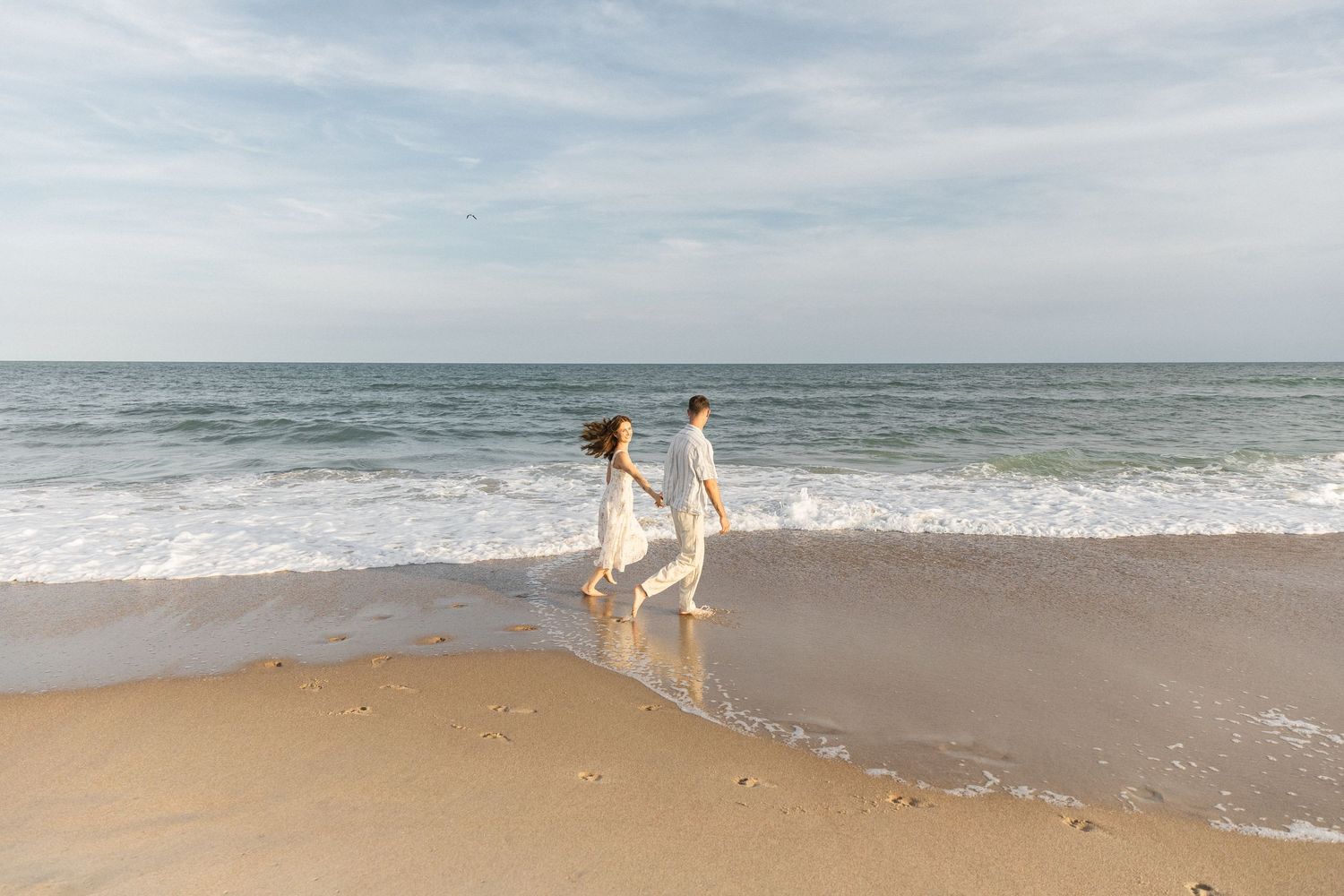 Couple running on the beach shoreline holding hands, laughing and having fun during their coast NC photoshoot