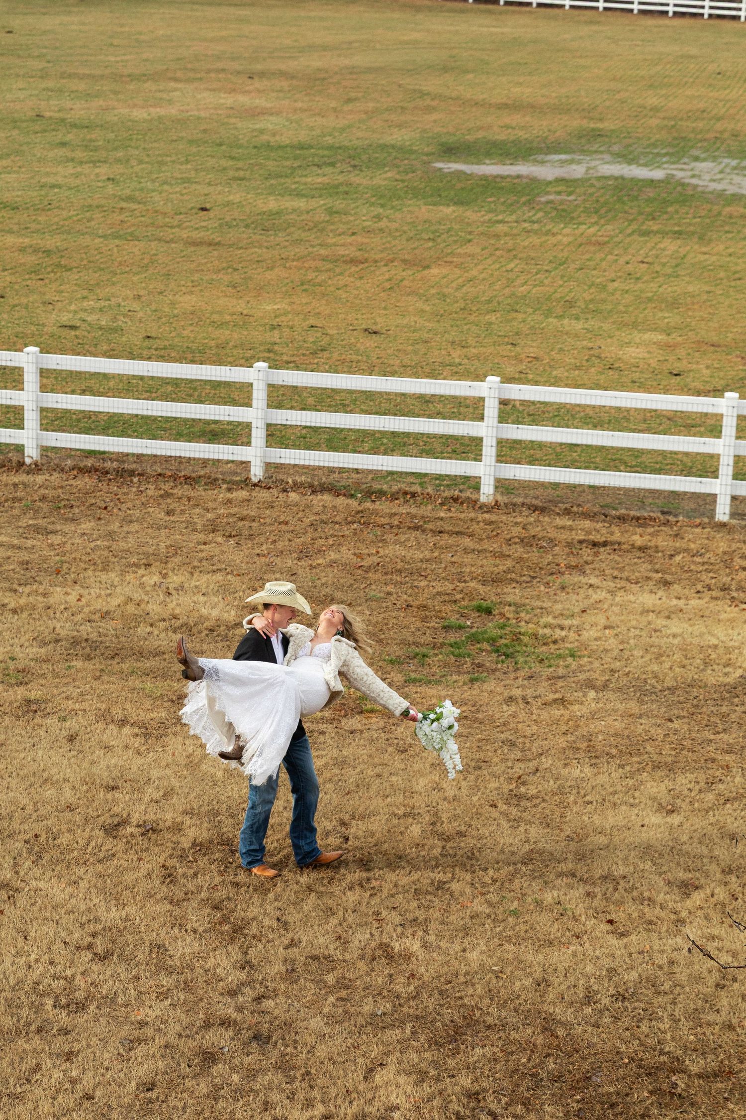 Rainy elopement in North Carolina. Groom in cowboy hat lifts bride in white dress in rustic farm field with white fence backdrop.