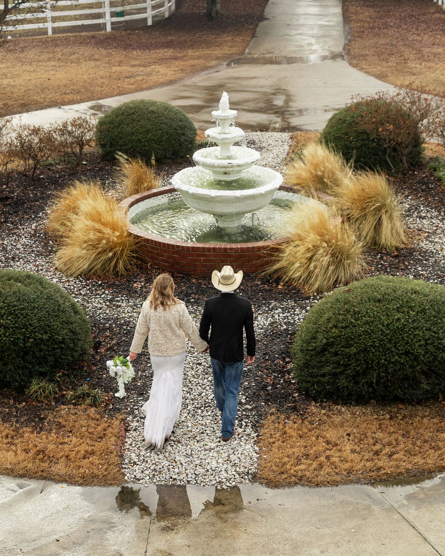Aerial view of a bride and groom walking toward a tiered garden fountain surrounded by trimmed hedges during their rainy day elopement in NC