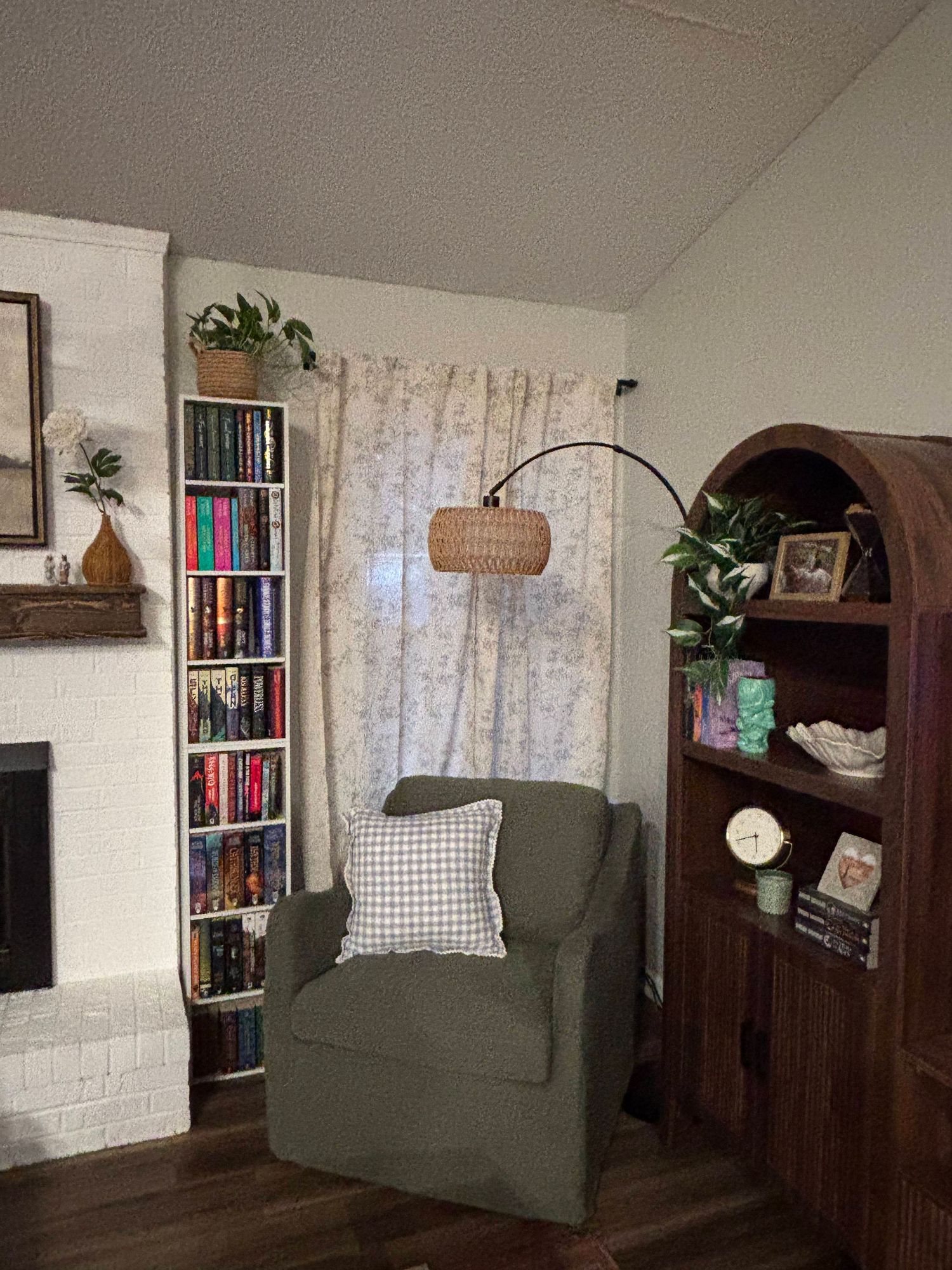 Cozy reading nook with green armchair, arc rattan lamp, tall bookshelf, wooden cabinet, and floral curtains in a warm living room.