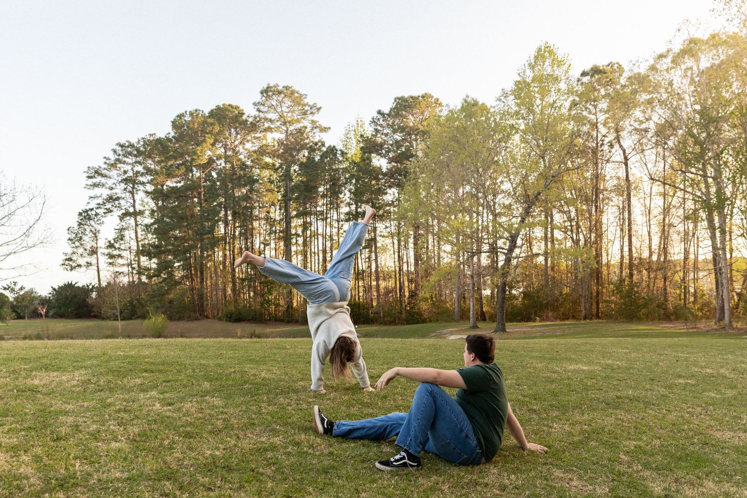 Person doing a cartwheel beside a friend sitting on grass in a sunny park with tall trees in the background.