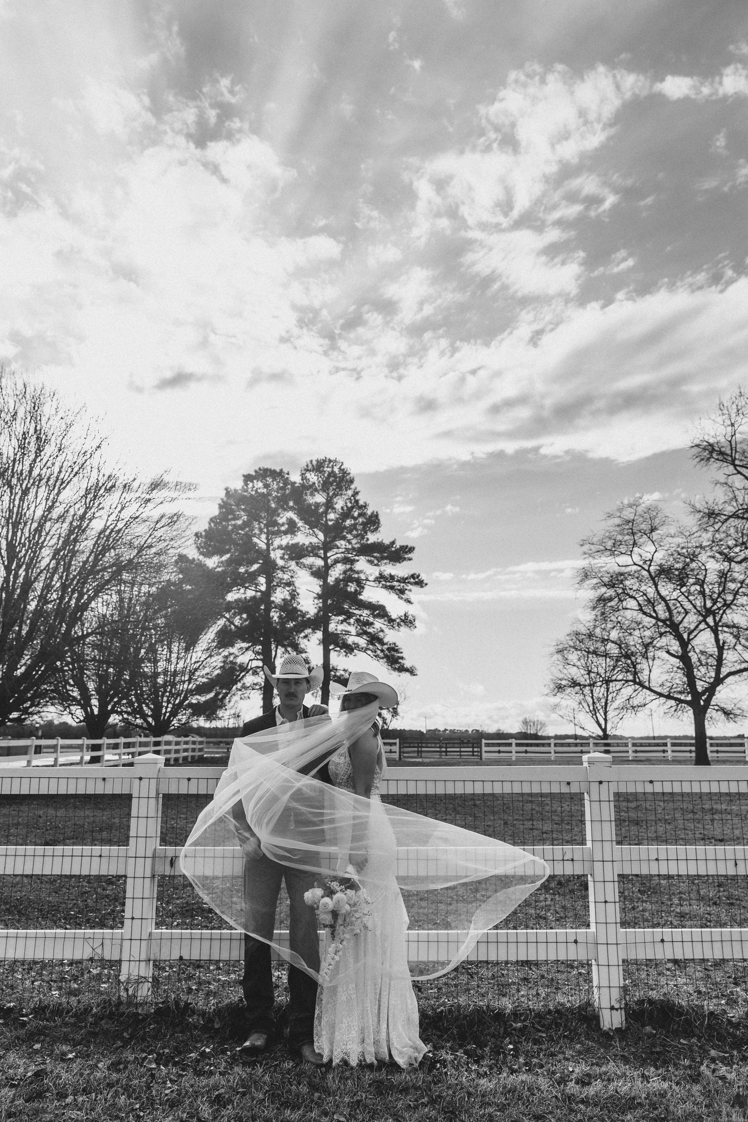 Black and white photo of a bride and groom kissing by a white fence, her veil flowing in the wind under a dramatic sky.
