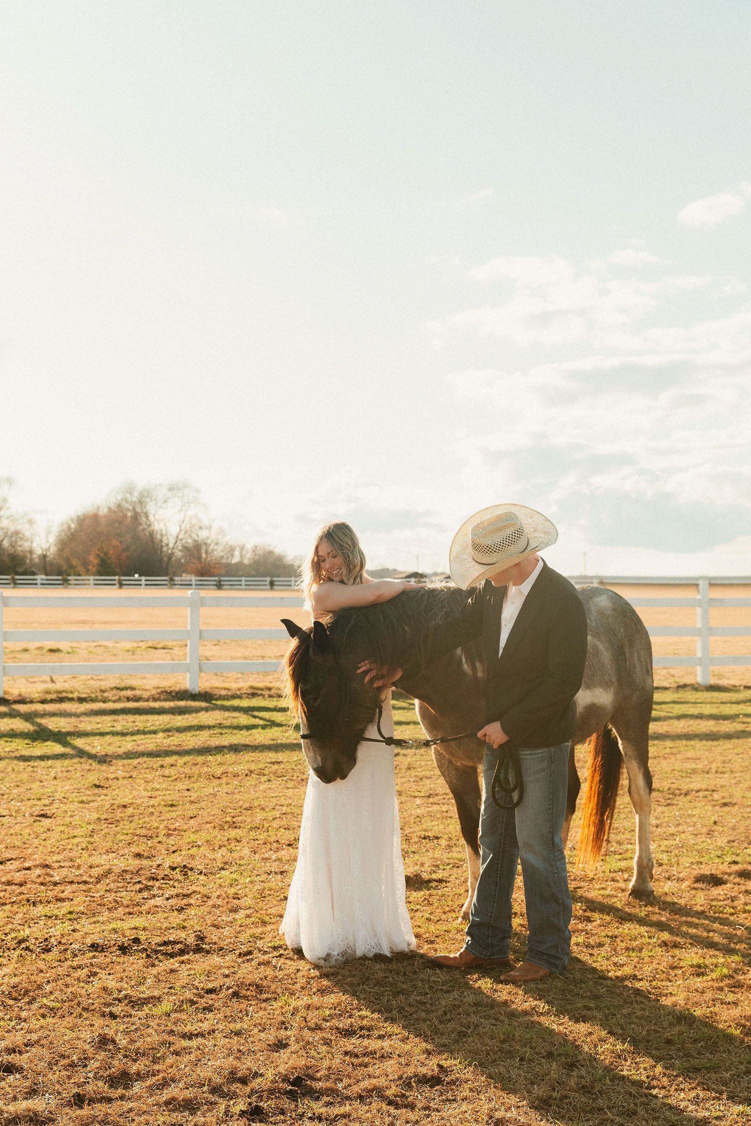 Bride and groom in cowboy hat petting a dark horse at golden hour on a sunny farm with white fence.