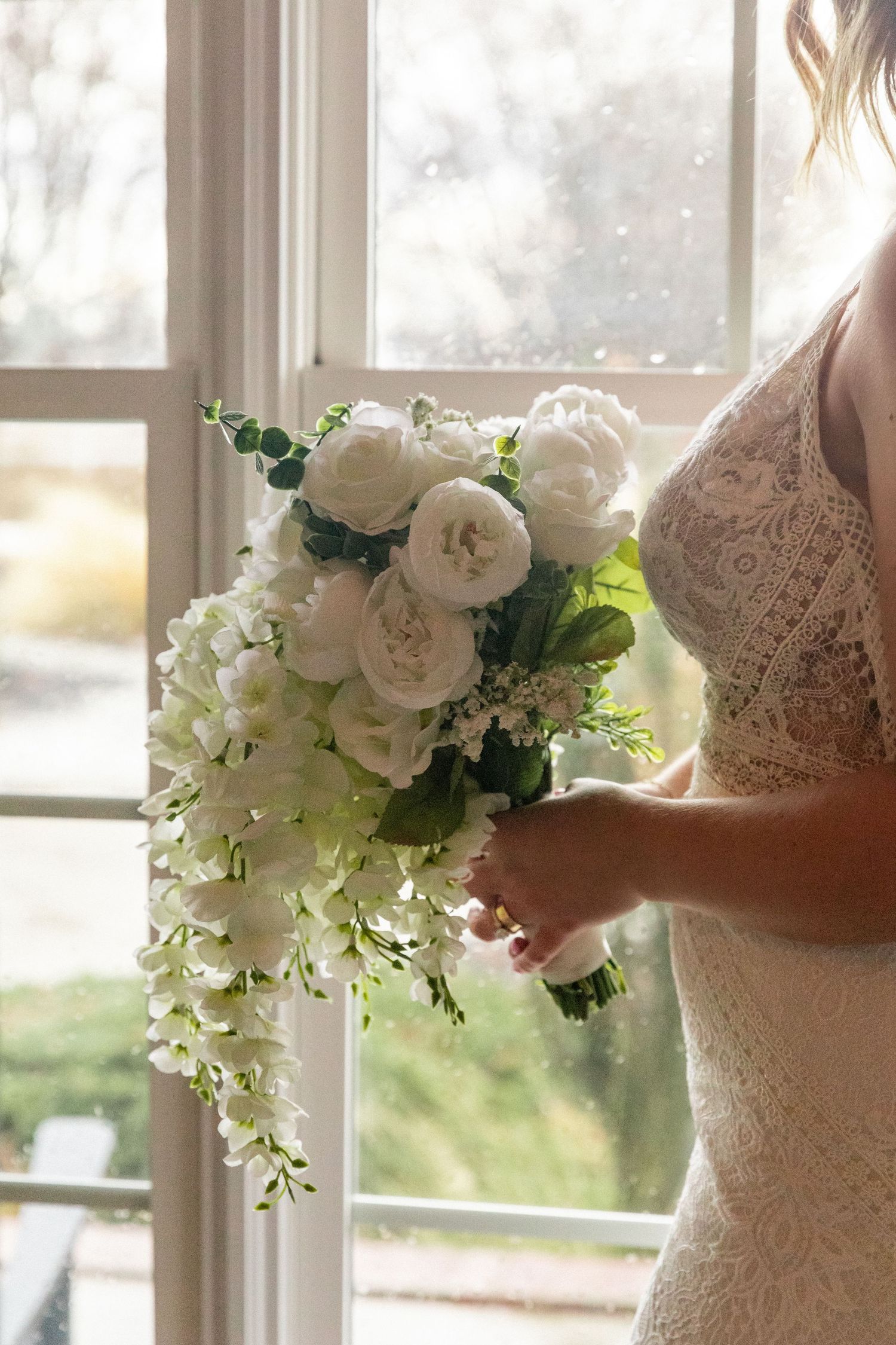 Bride holding a cascading white wedding bouquet with peonies and hydrangeas, standing by a sunlit window in a lace gown.