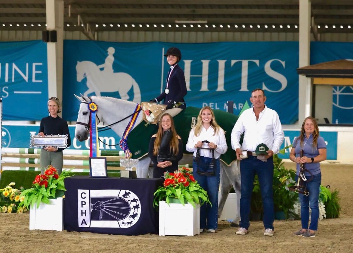 Winner's circle at HITS equestrian show with rider on white horse surrounded by five people holding awards.
