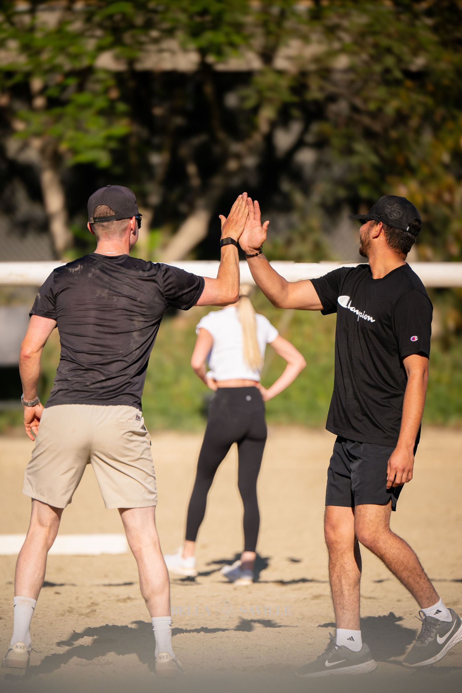 Two men high-five on sandy volleyball court with a woman standing in the background.
