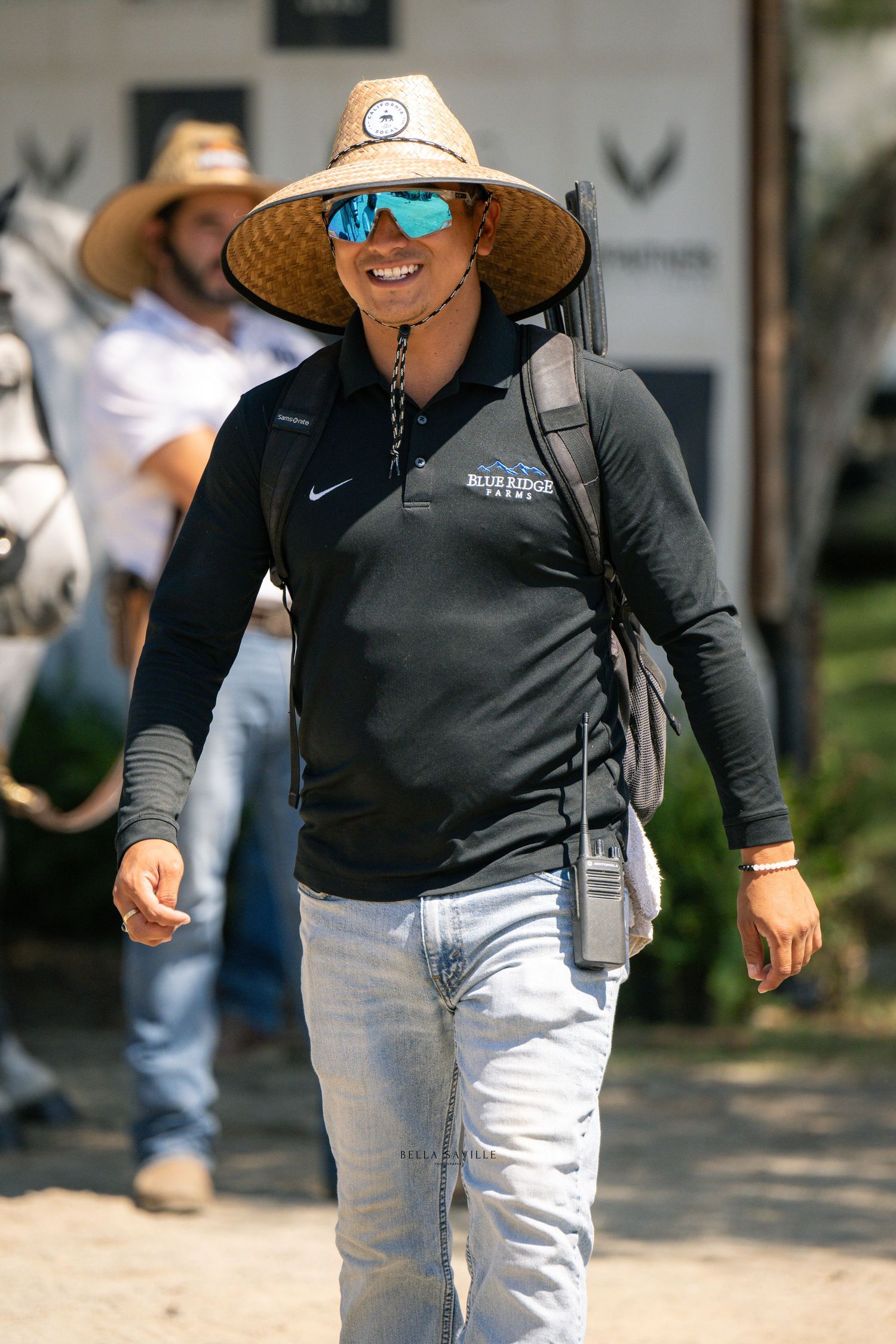Smiling woman in Blue Ridge Farms polo, straw hat, and blue sunglasses walks at horse show.