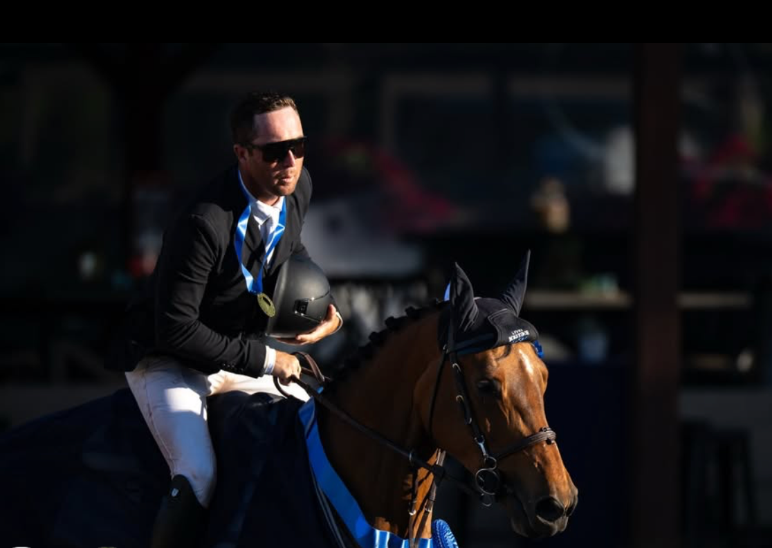 Sean Leckie wins 1.35 Welcome. Equestrian rider in black jacket rides bay horse decorated with blue ribbon at competition.