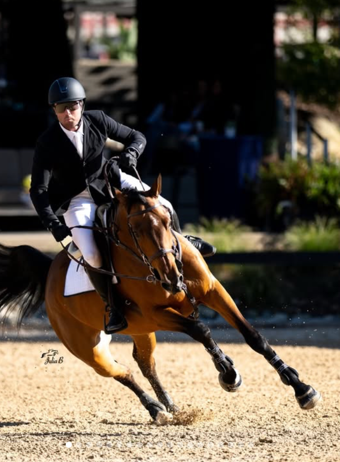 Equestrian rider in black jacket and helmet on a bay horse competing in a show jumping event on a sandy arena. Sean Leckie wins 1.35 Welcome