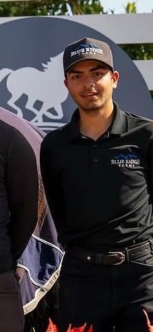 Man wearing Blue Ridge Farms black polo shirt and matching cap standing outdoors near a horse farm sign.