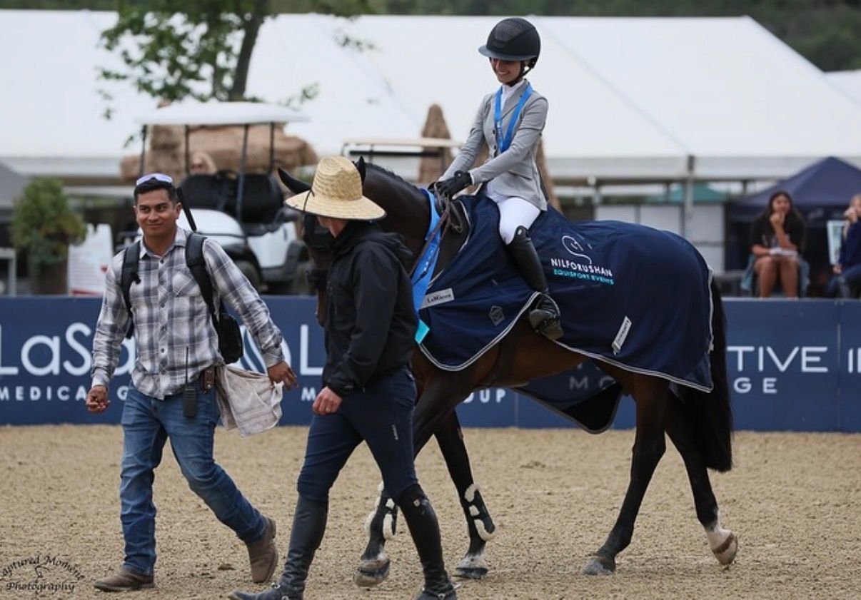 Young equestrian rider on dark horse with blue blanket, led by handlers at outdoor show jumping competition.