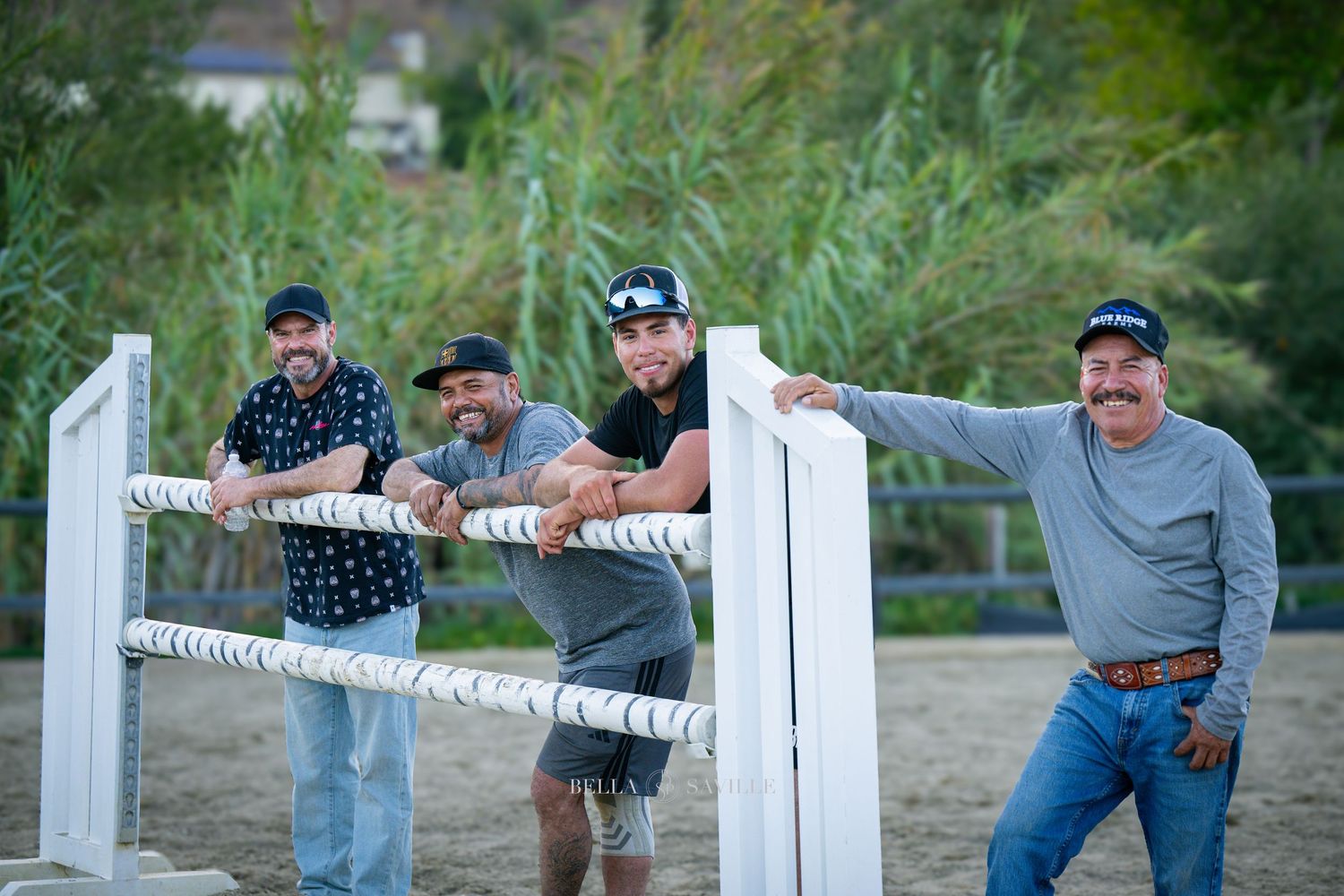 Four men pose together holding white fence rails at an outdoor equestrian arena at sunset.
