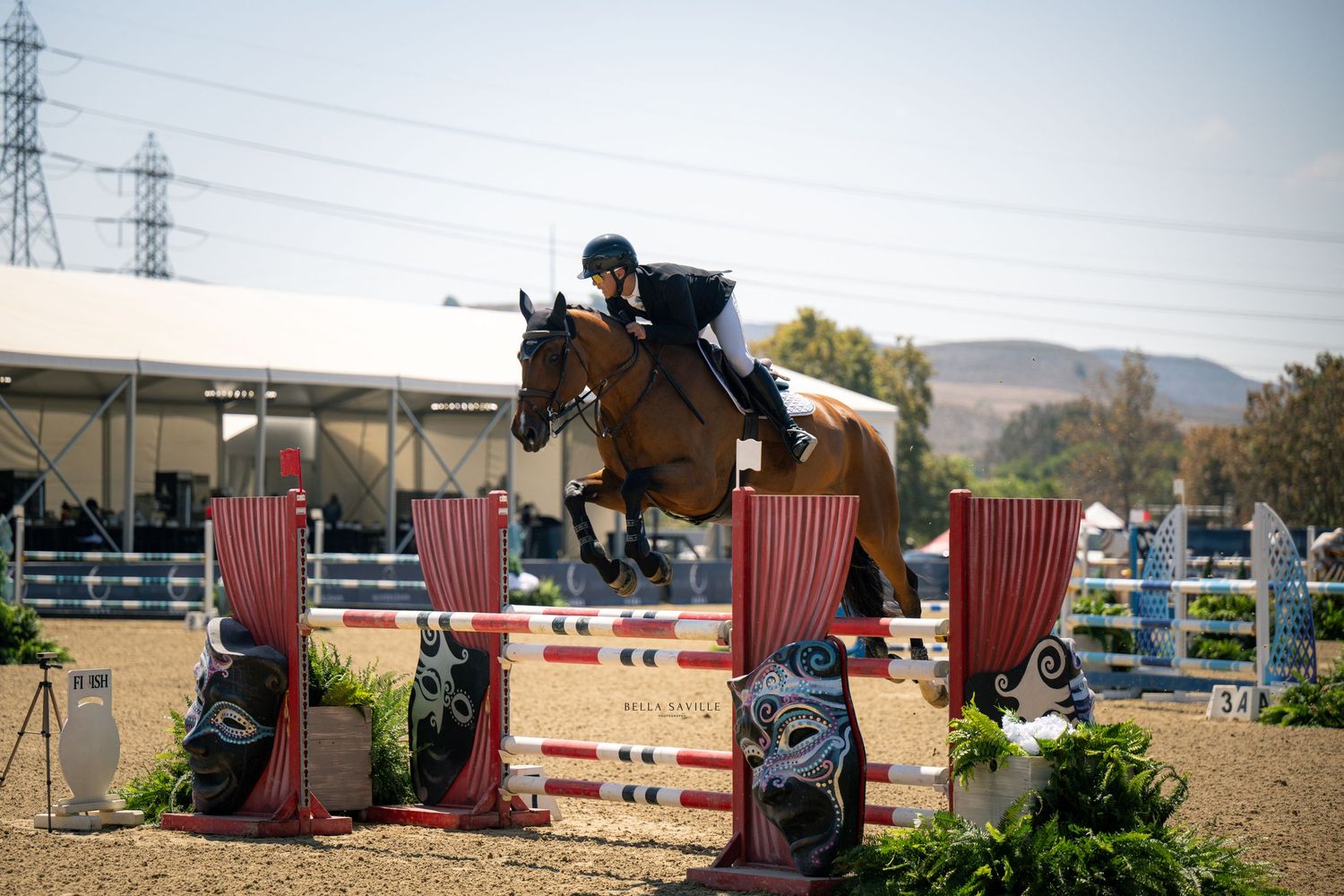 Equestrian rider on chestnut horse soars over colorful jump at outdoor show jumping competition.