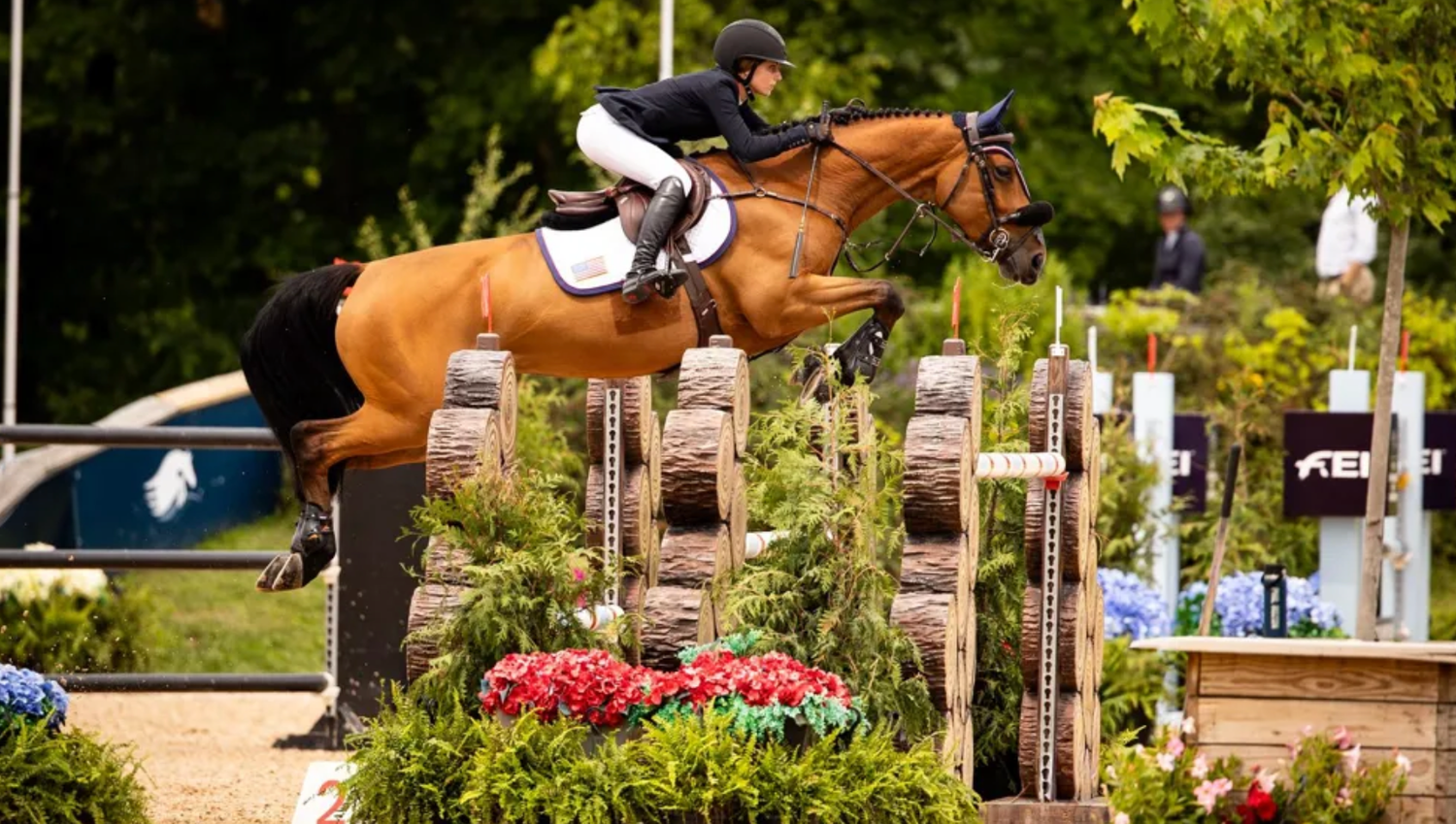 Equestrian rider on chestnut horse jumping over colorful flower obstacle in outdoor show jumping competition.