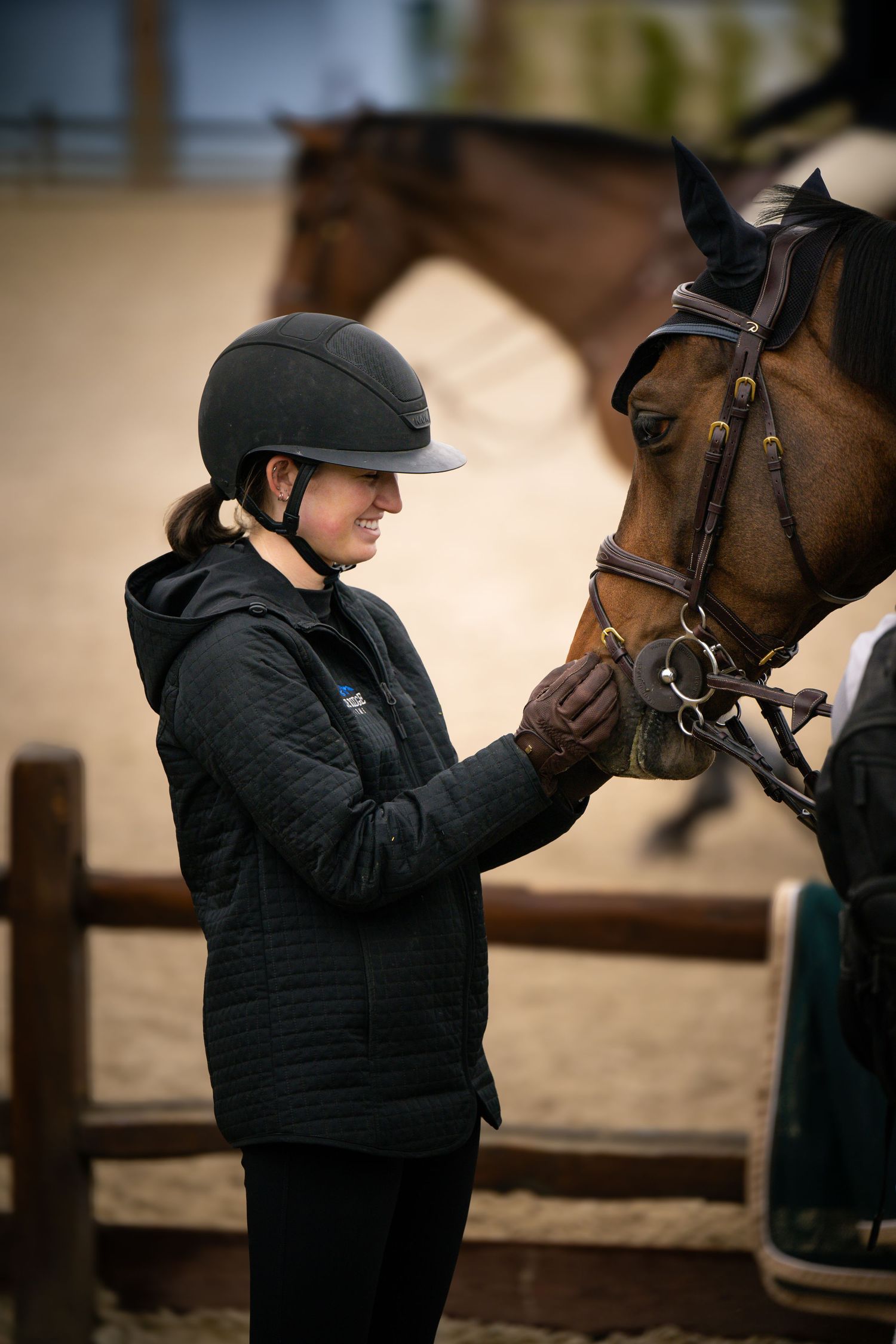 Female equestrian in black helmet adjusting bridle on a bay horse in indoor arena.