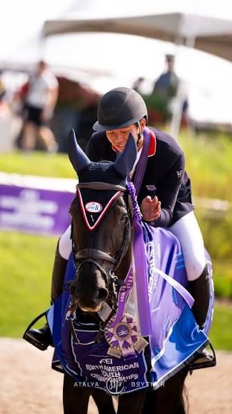Equestrian rider in black helmet celebrates FEI North American Youth Championships win on decorated horse with purple ribbon.