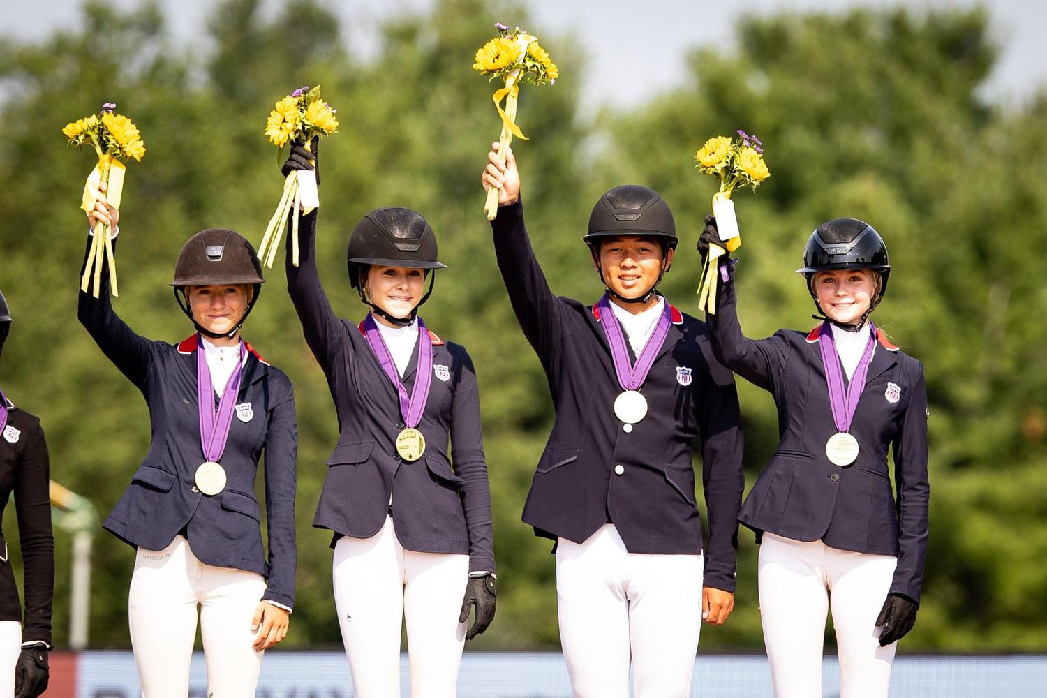 Four equestrian riders in navy jackets celebrate on podium, holding yellow flowers and medals at a competitive horse show event.