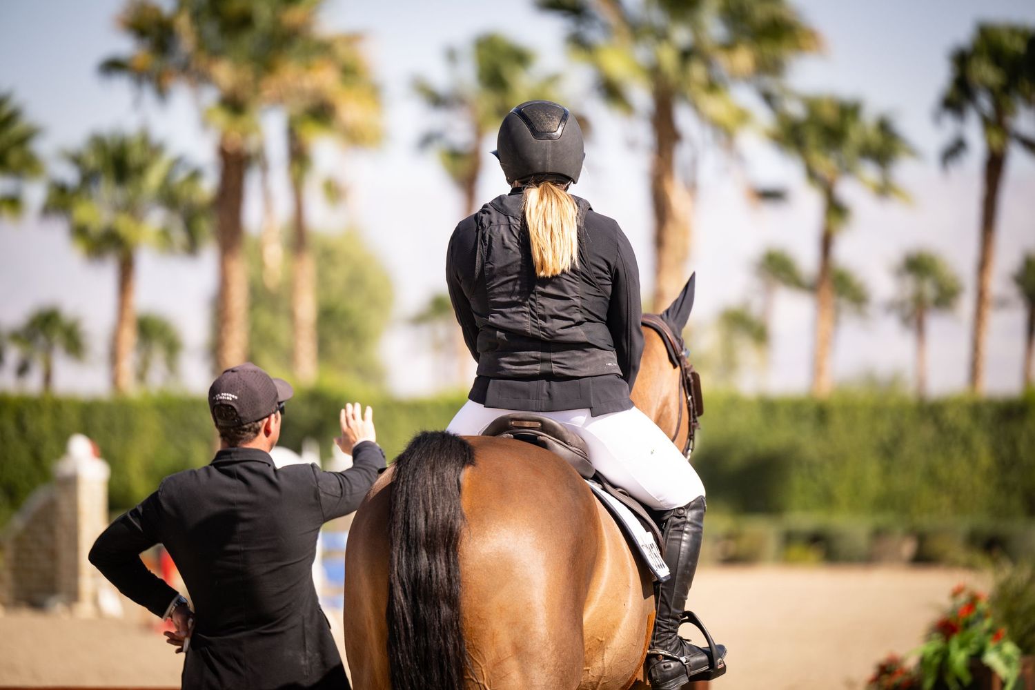 Blonde female rider on bay horse receiving instruction at palm tree-lined equestrian venue.