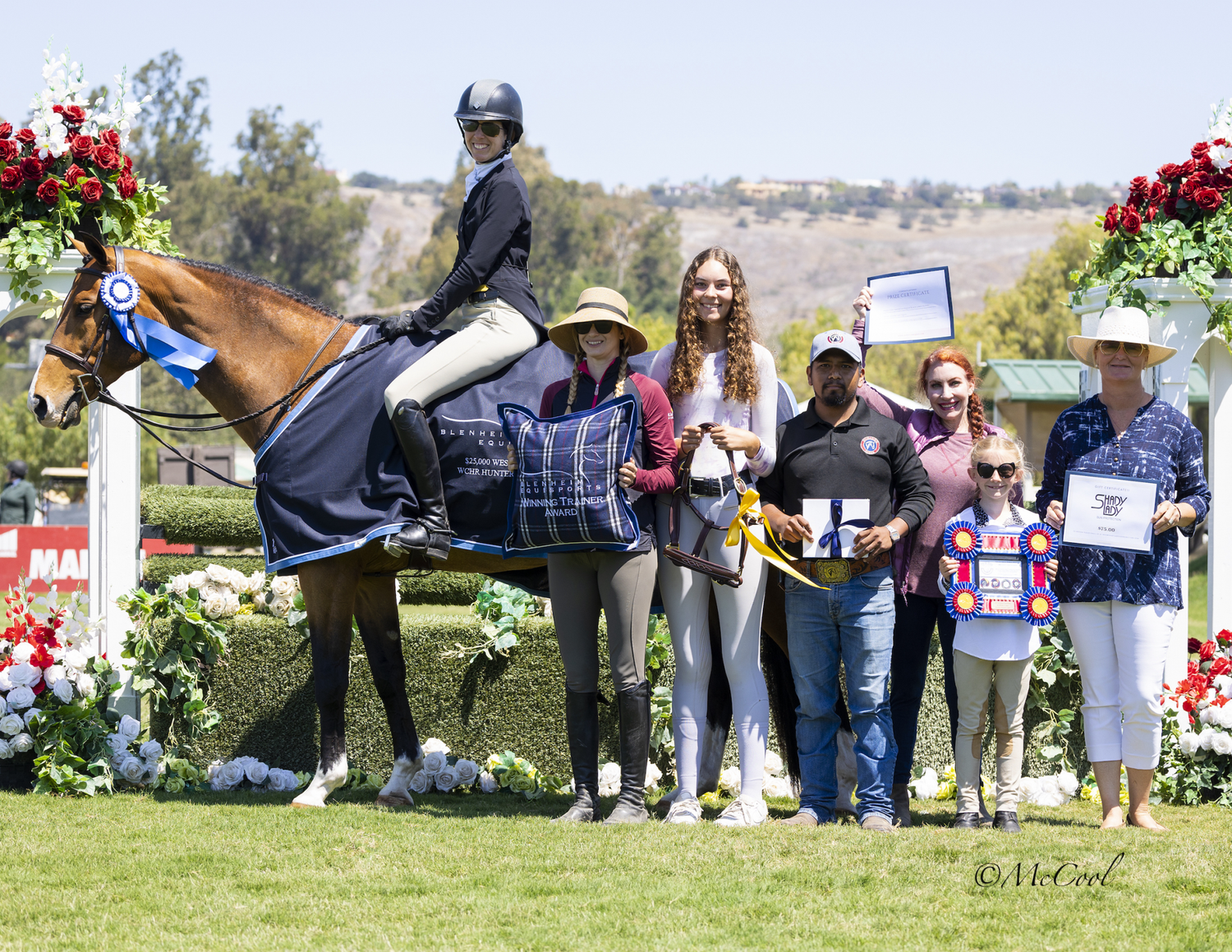 Award ceremony with equestrian rider on horse surrounded by smiling presenters at outdoor show.