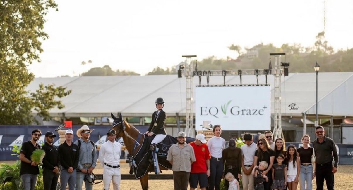Equestrian rider on horseback at EQGraze show jumping competition with crowd of spectators gathered around.