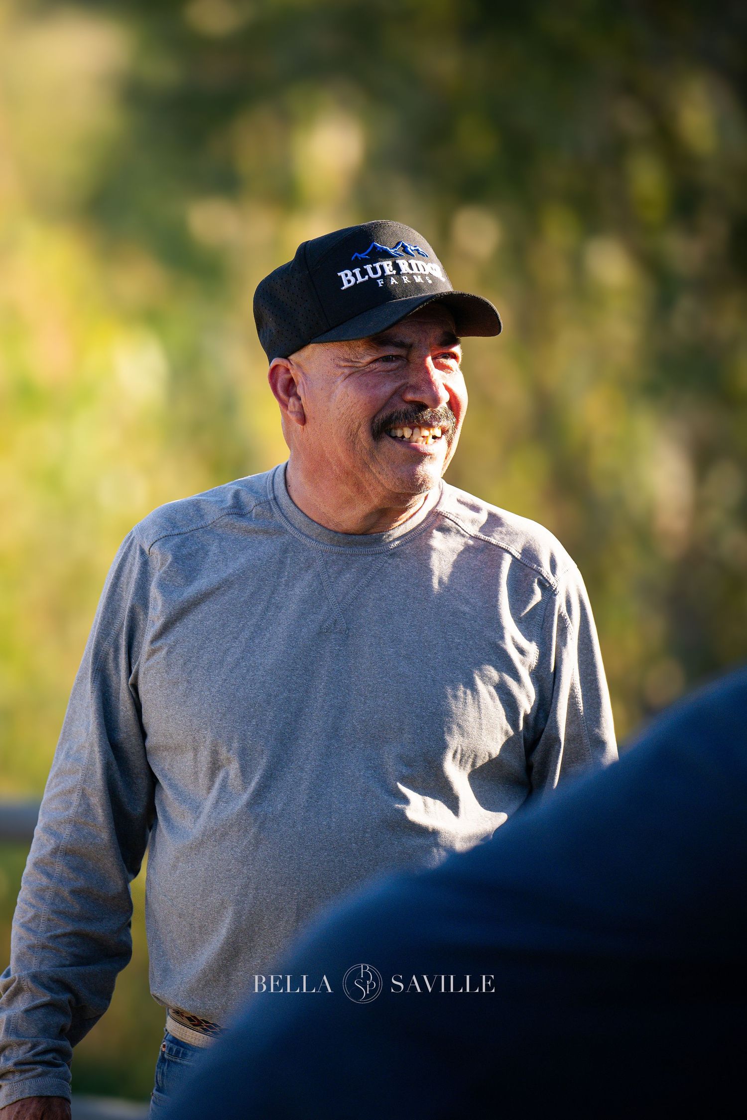 Smiling middle-aged man in Blue Ridge Farms cap enjoys outdoor equestrian event in warm light.