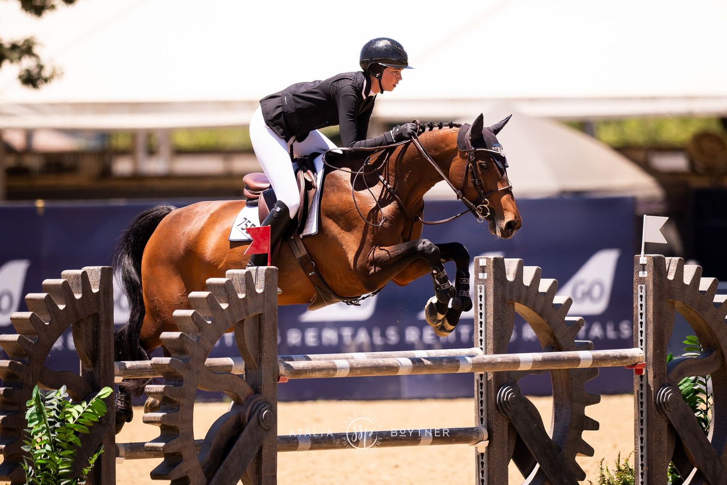 Equestrian show jumper on bay horse clearing metal fence obstacle at outdoor competition with sponsors.