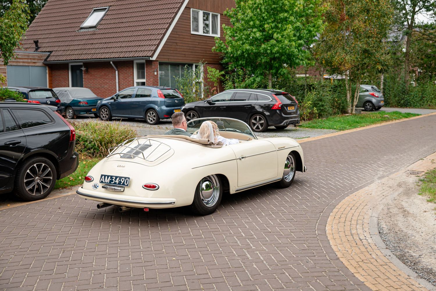 Vintage cream Porsche 356 speedster convertible parked on brick road in residential neighborhood.