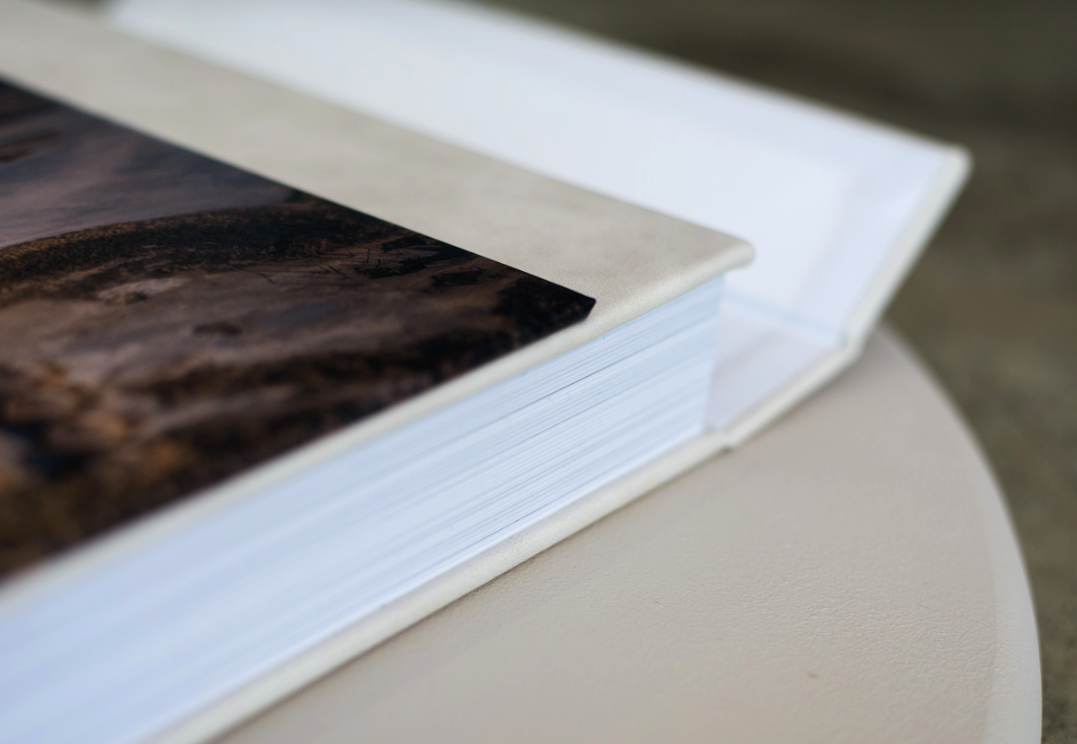 Close-up of a luxury photo album with a landscape image cover and thick white pages resting on a round cream-colored surface.