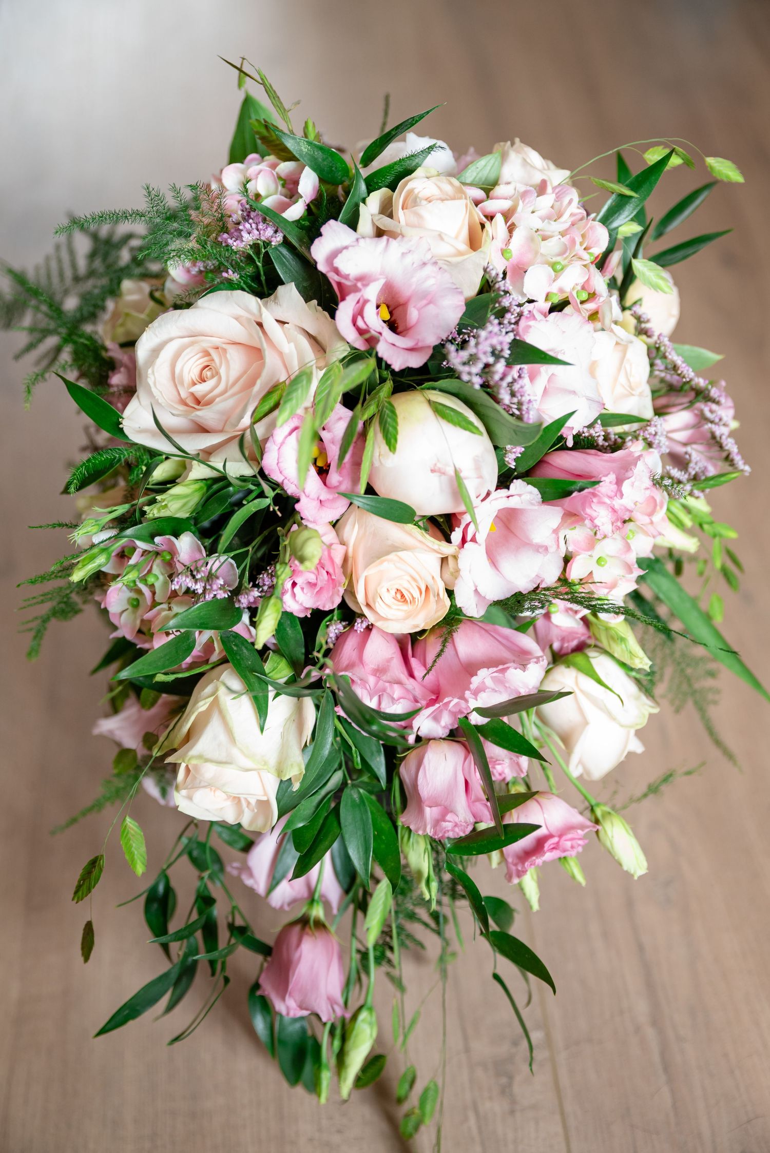 An elegant cascading bridal bouquet of pink roses, peonies, and greenery on a wooden surface.