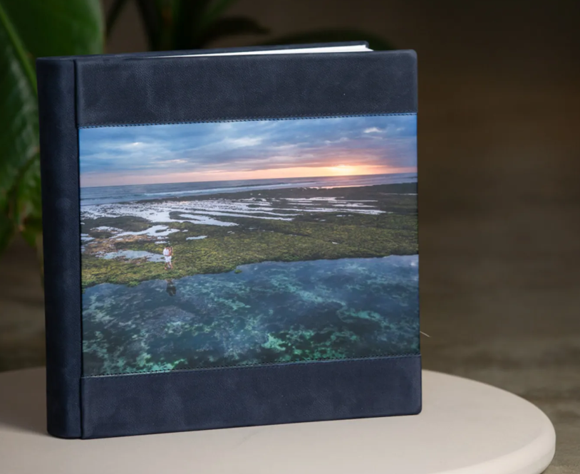 Navy blue photo album with coastal sunset and reef cover image displayed on a white table.