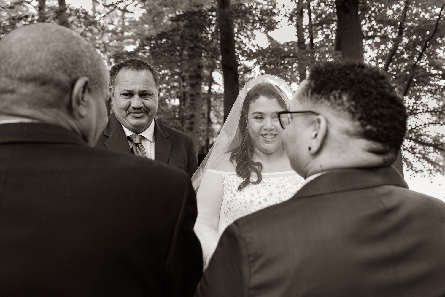Black and white wedding ceremony photo showing bride and groom with officiant outdoors among trees.