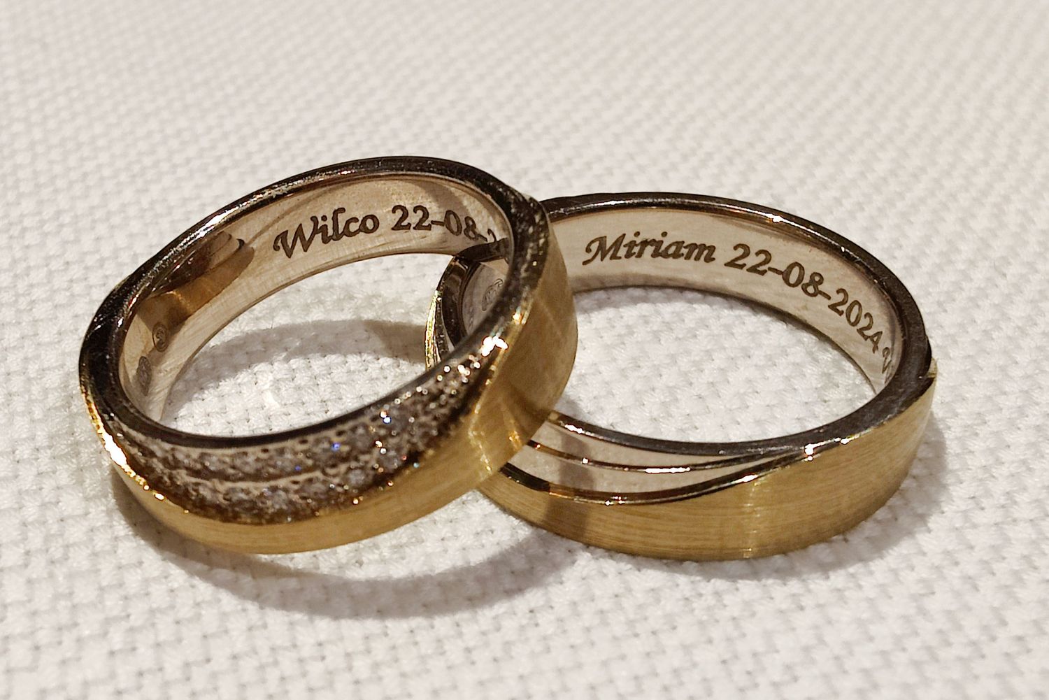 Two gold wedding bands engraved with names Wilco and Miriam and date 22-08-2024 on white fabric.