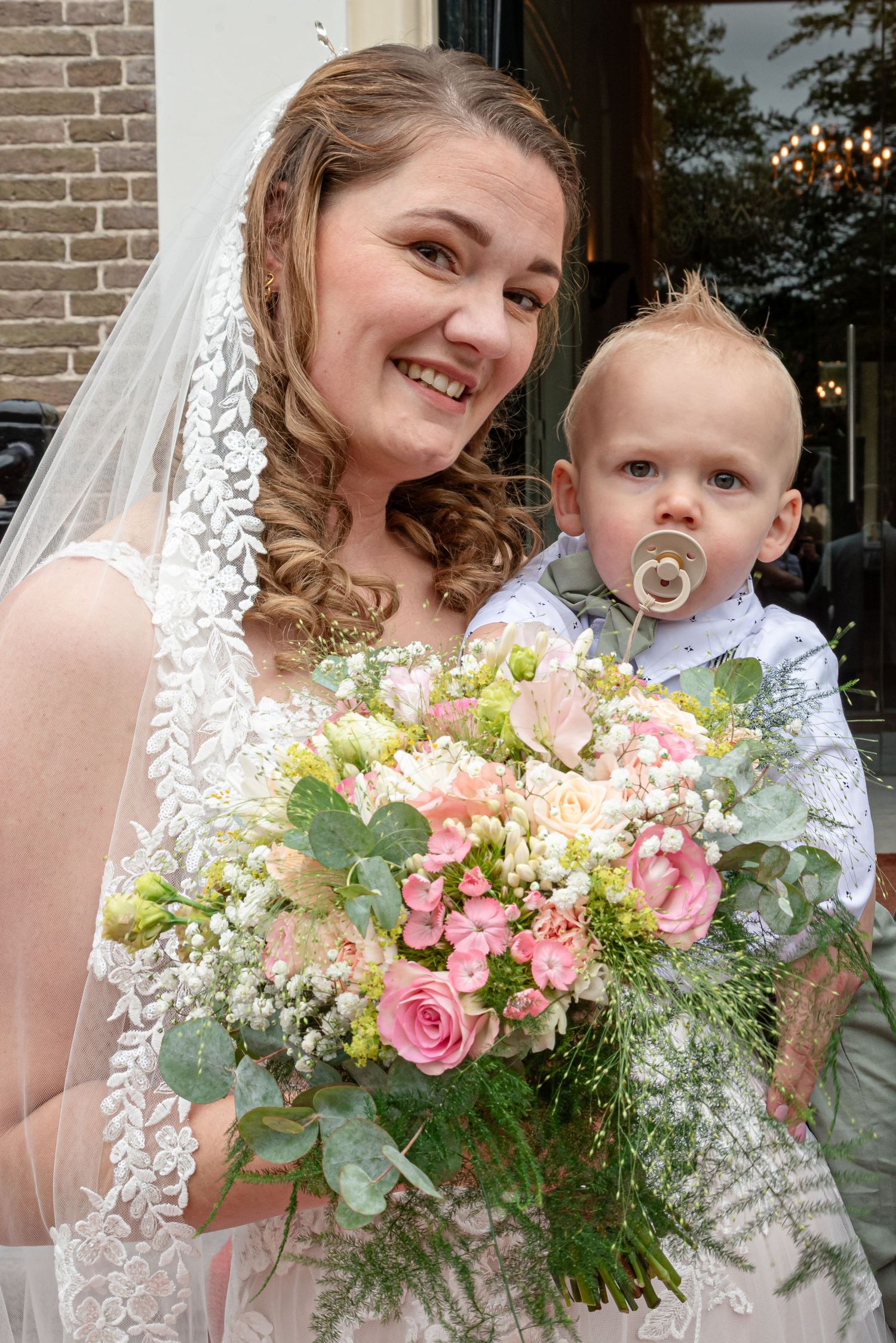 A smiling bride in a lace veil holds a pink and white bouquet while carrying a baby with a pacifier.