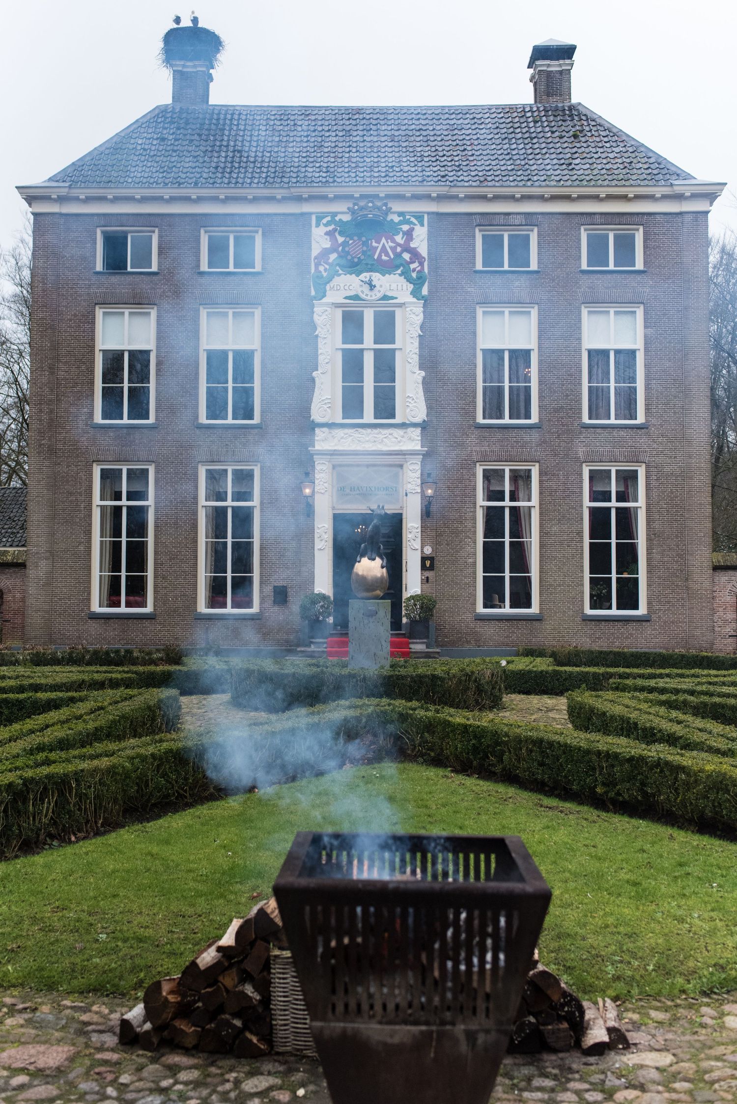 Historic Dutch manor house with formal hedge garden and smoking fire basket in foreground on overcast day.