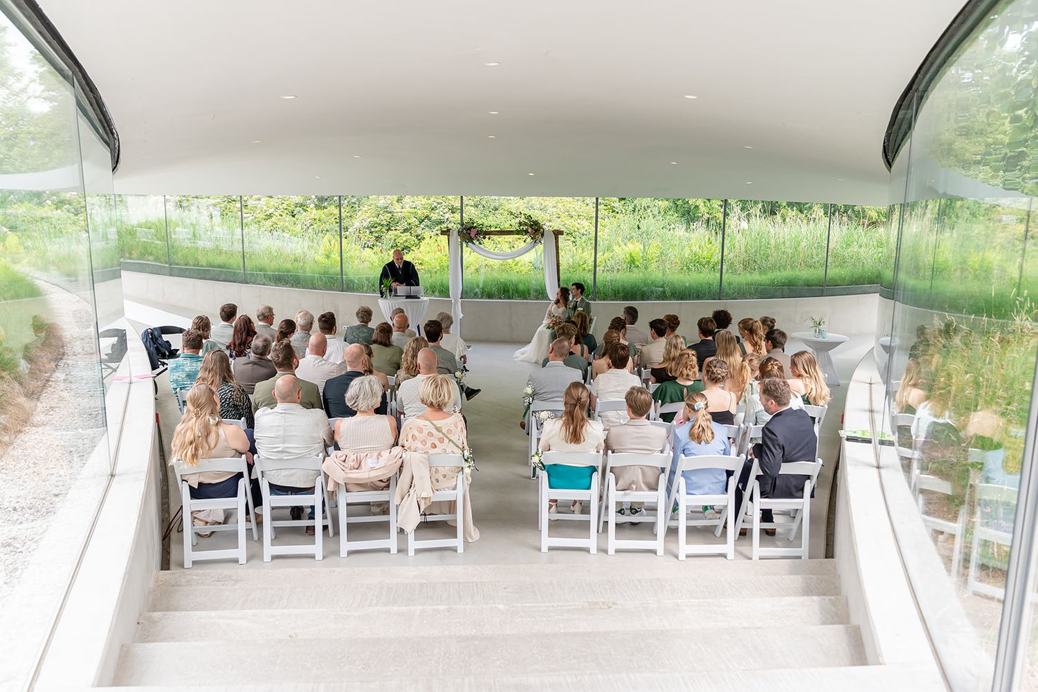 Outdoor wedding ceremony with guests seated in white chairs under a modern glass pavilion surrounded by lush greenery.