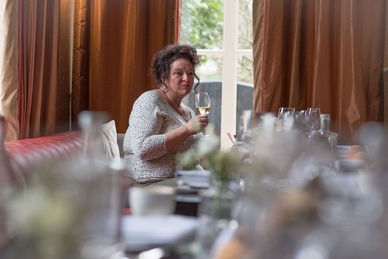 Woman in lace top holding wine glass at elegant dinner table with floral centerpieces and warm curtains.