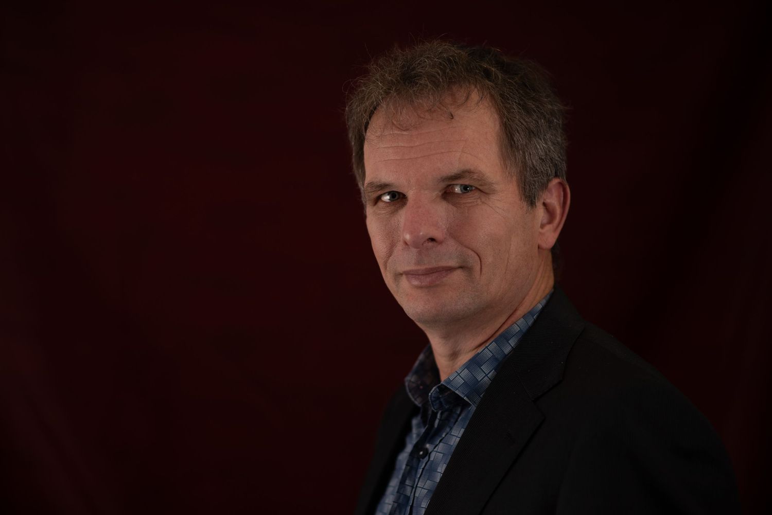 Middle-aged man in dark blazer poses against deep red background in professional portrait photography.