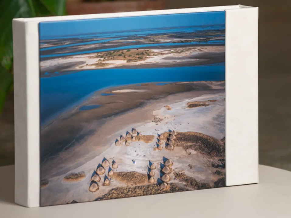 White hardcover photo book displaying an aerial image of winding blue waterways and sandy islands with small village huts on the shore.