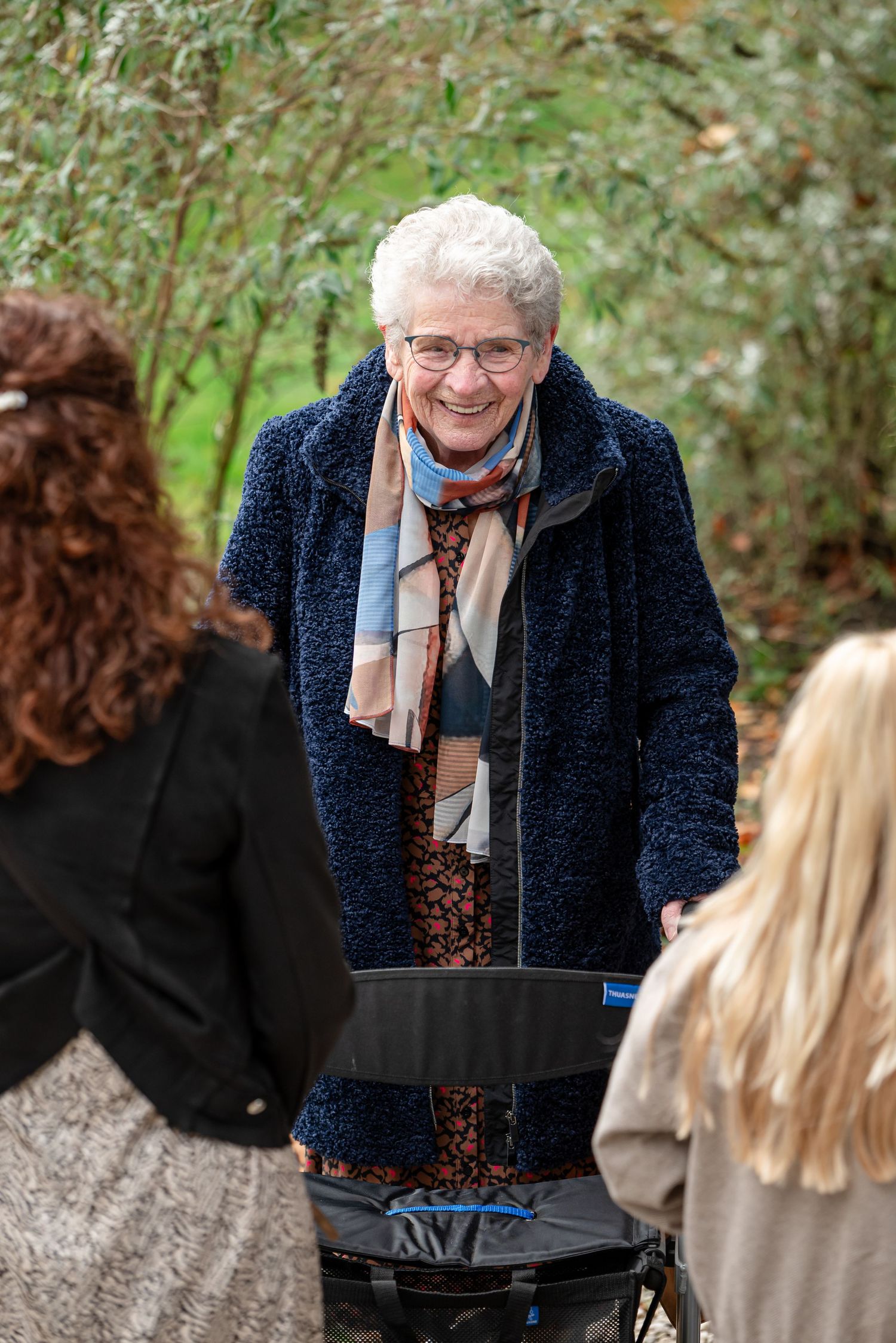 Oudere vrouw met een rollator in een nette jas en sjaal. Ze is zichtbaar tussen andere mensen door. Voorbeeld van een kostbare foto van Oma.