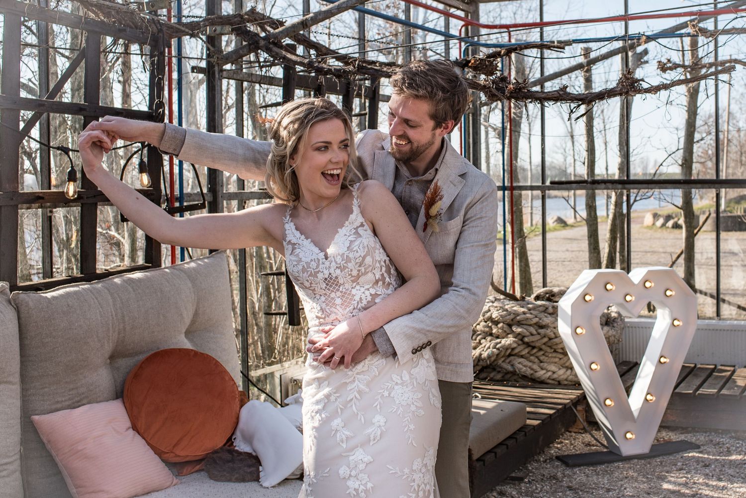 Bruidspaar danst lachend in een kas voor een hart-lamp in het vroege voorjaar. Buiten is het nog kaal maar de zon schijnt naar binnen.